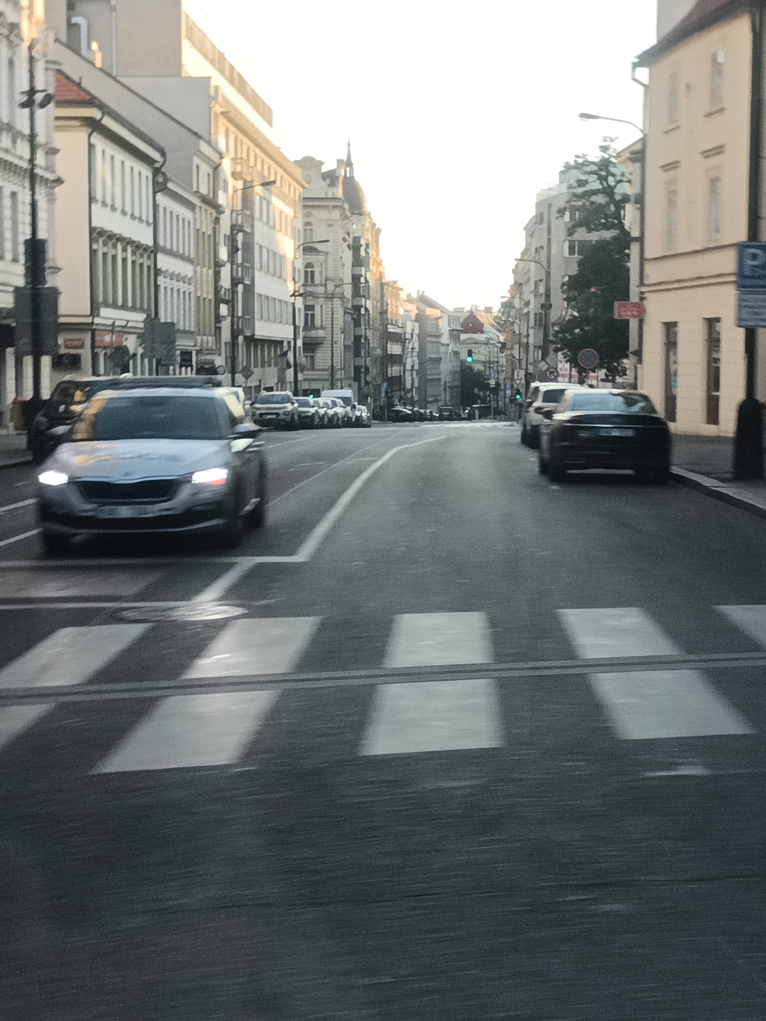 An eye-level, medium shot captures a street scene in Prague, Czechia, with a noticeable blur indicating motion. The perspective is from within a moving vehicle, looking forward down a paved road lined with tall, multi-story buildings on both sides. The architecture suggests a European urban environment, with a mix of styles, some featuring decorative elements. The sky is bright and overcast, with the sun likely low on the horizon, casting a soft light.
In the foreground, a pedestrian crossing is visible, its white stripes blurred by movement. The primary subject is a silver car approaching the crossing from the left, its headlights illuminated. The car appears to be a sedan or hatchback. Further down the road, several other cars are visible, some parked, others in motion, forming a line. A traffic light, showing green, is visible in the distance. Street lamps are present along the sidewalks.
The overall impression is that of a typical urban commute in Prague during daylight hours, possibly during the morning or evening rush. The motion blur lends a dynamic quality to the image, emphasizing the passage of time and the ongoing activity of the city.
