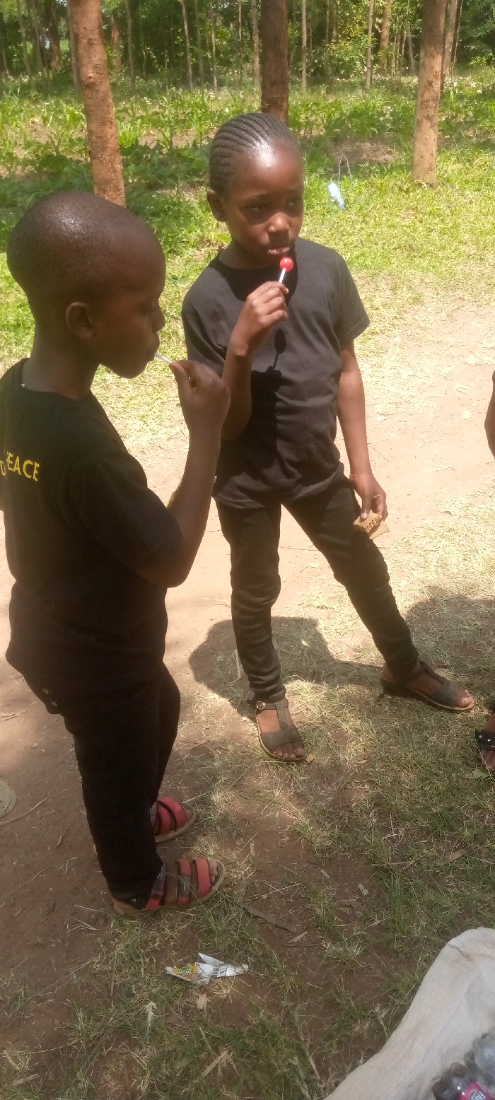Two young boys stand in a grassy clearing in the forest. The boy on the left is wearing a black shirt with "PEACE" written on it and black shorts. He is wearing red sandals. The boy on the right is wearing a black shirt and jeans, and brown sandals. He is holding a red lollipop in his right hand. The boys are standing on a dirt path. The forest is in the background. There is a tree on the left side of the image. The location is a forest clearing in Lwandeti, Kenya, near the coordinates (latitude: 0.579, longitude: 34.808).