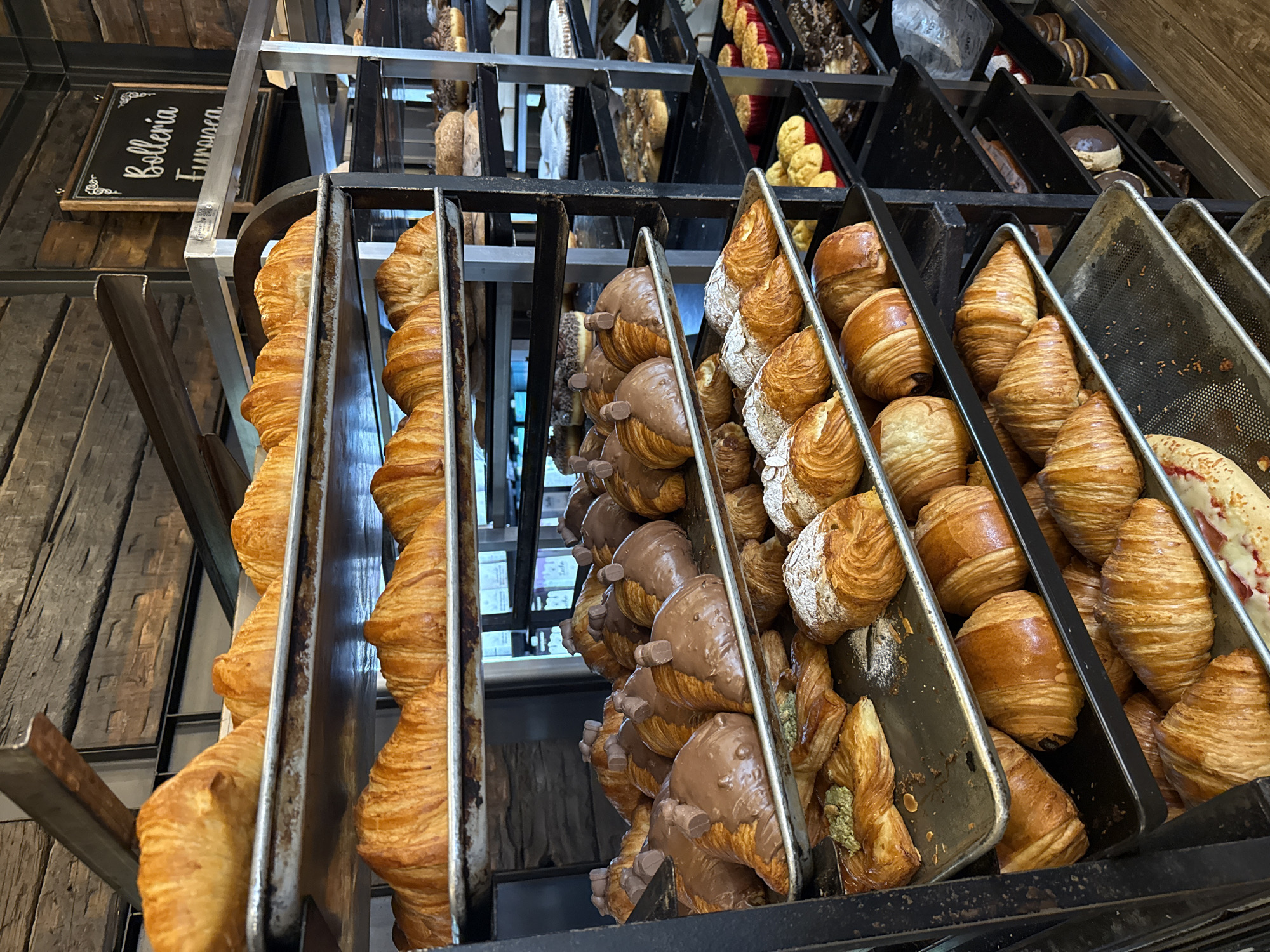 The image depicts a bakery display case filled with various types of croissants. The croissants are arranged in metal trays. Some are plain, others are covered in chocolate and topped with small chocolate pieces, while others are sprinkled with powdered sugar or filled with pistachio cream. The display case is made of black metal and has a sign that reads "Bolleria Europea". The location is a bakery in Puebla, Mexico.