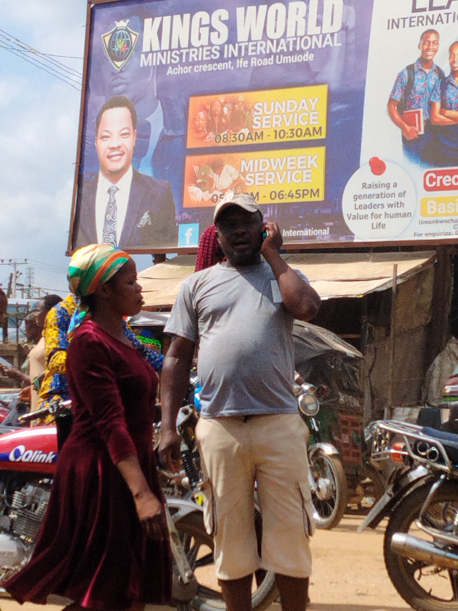 The image depicts a street scene in Umuode, Nigeria. A large billboard advertises "Kings World Ministries International" with service times displayed. In the foreground, a woman in a red dress and a headscarf stands next to a man wearing a grey t-shirt, a cap, and khaki shorts. The man is talking on a cell phone. Several motorcycles are parked nearby.