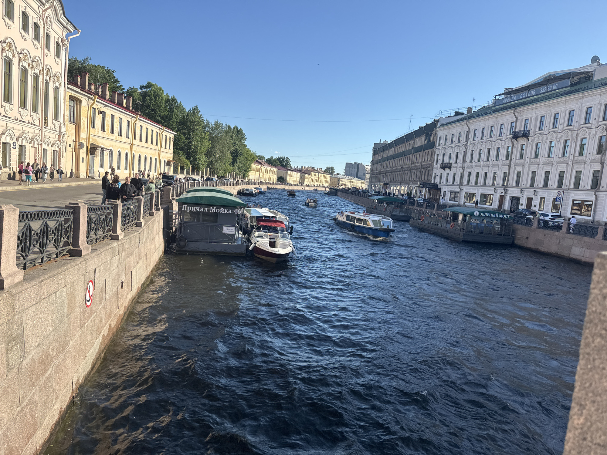 This is a medium-distance, eye-level shot of the Moika River in Saint Petersburg, Russia. The water is a deep, dark blue with moderate ripples. Several boats are visible, including tour boats docked at "Причал Мойка 46" (Moika Pier 46) and another labeled "ЗЕЛЁНЫЙ МОСТ" (Green Bridge). A few smaller motorboats are also navigating the river.
The left bank of the river features a light yellow, ornate building with multiple arched windows and a balustrade running along the river. A stone embankment with a decorative metal fence lines this bank, and several pedestrians are walking along the sidewalk. A red and white circular sign with a red anchor crossed out is affixed to the embankment wall.
The right bank is dominated by a long, pale, classical building with numerous windows and balconies. Further down the river, more buildings and trees line the banks. The sky is a clear, bright blue, indicating a sunny day. The overall impression is of a vibrant city scene with a busy waterway.
