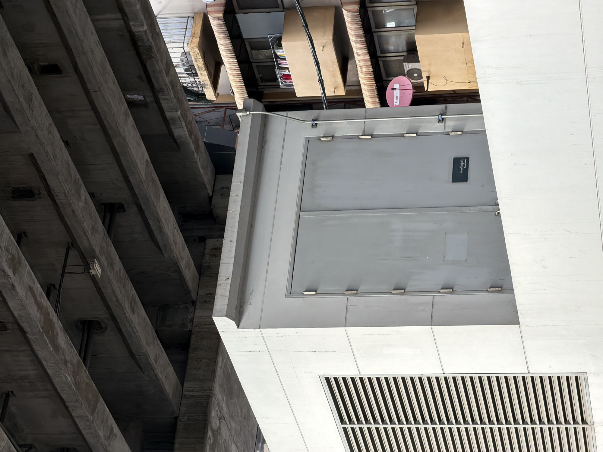 The image shows a concrete structure with large double doors and a louvered panel, situated beneath a large overpass. To the right, a multi-story building with balconies and awnings is visible, featuring clothes hanging on a line and satellite dishes on the exterior. The scene is set in an urban environment, likely in Bangkok, Thailand, given the visible signage that appears to be in Thai. The overall impression is one of urban infrastructure and everyday life coexisting.