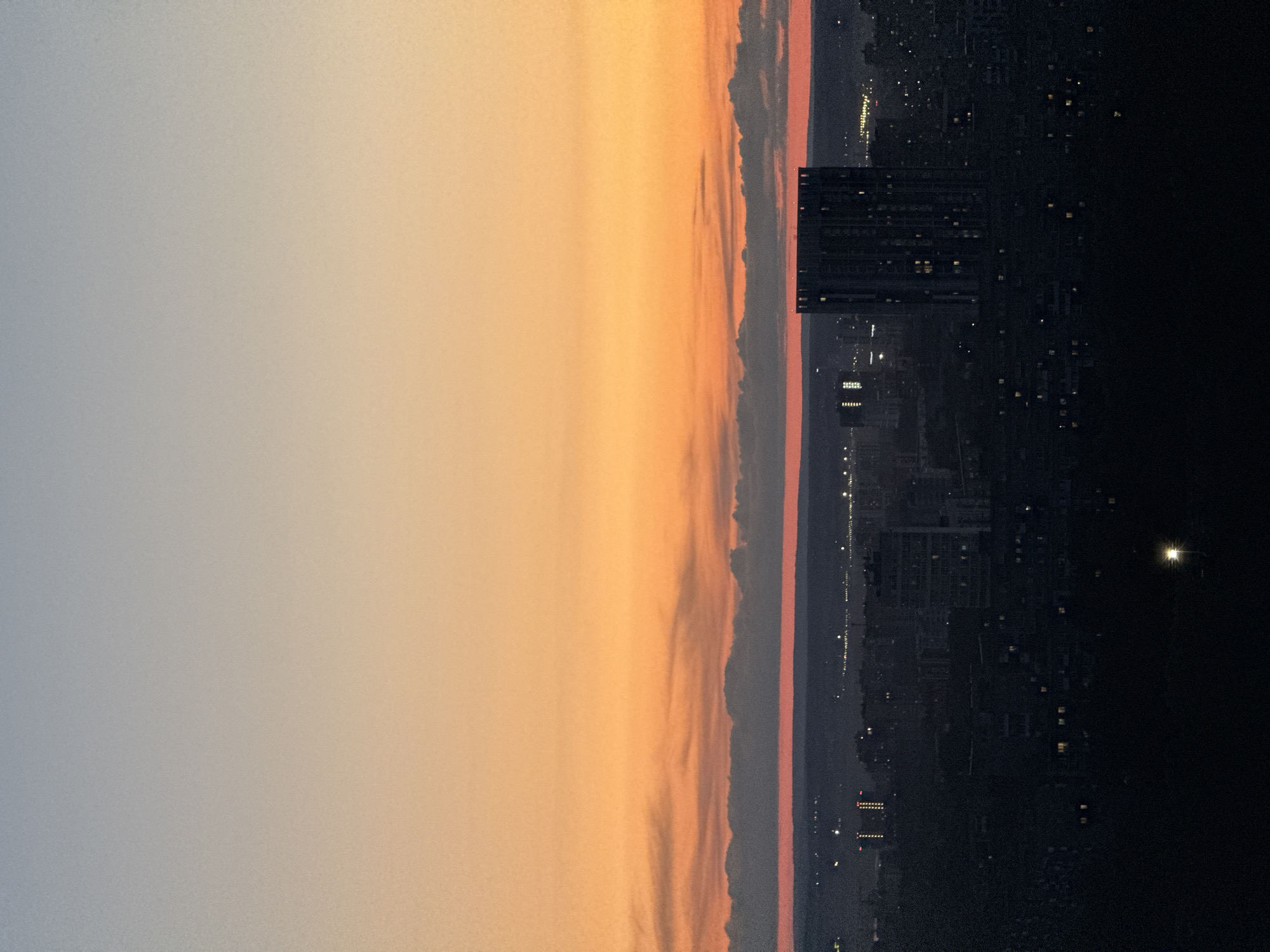 The image presents a wide, panoramic view of a city landscape during either dusk or dawn. The upper portion of the frame features a sky transitioning from a soft, light grey-blue on the left to a warm, glowing orange and yellow towards the right, suggesting the position of the sun. Below this gradient sky, a distinct, elongated band of clouds, infused with pink and orange hues, stretches horizontally across the mid-ground, creating a natural horizon line.
Below the cloud band, the city unfolds in deepening darkness. Numerous scattered lights from buildings and streets are visible across the urban expanse, beginning to illuminate the night or fade with the rising sun. Several large, dark high-rise buildings are discernible, with one prominent structure on the right side of the frame featuring multiple illuminated windows. Towards the far right, a solitary, bright point of light, possibly a streetlamp or building light, stands out against the dim backdrop.
No people are visible in the scene. The image captures the serene transition between day and night over an urban environment. The overall mood is quiet and expansive, highlighting the natural beauty of the sky contrasting with the structured elements of the city. The photograph displays noticeable digital noise, particularly in the lighter, uniform areas of the sky and the darker city sections. There is no visible text. The setting is consistent with an urban view, such as one might find in Yug, Russia.