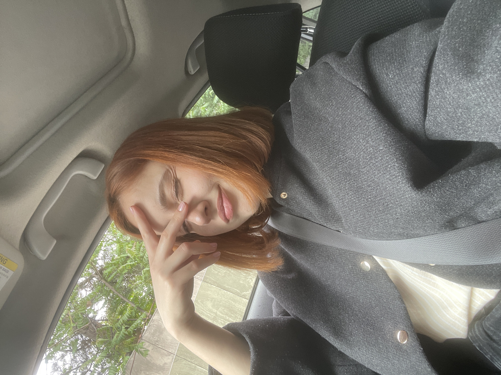 A young woman with red hair is sitting in the passenger seat of a car. She is wearing a dark gray coat over a light-colored striped shirt. The woman has her eyes closed and her hand resting on her forehead. A seatbelt is visible across her torso. Outside the car window, green trees and a gray building can be seen. The scene is likely in Tbilisi, Georgia.