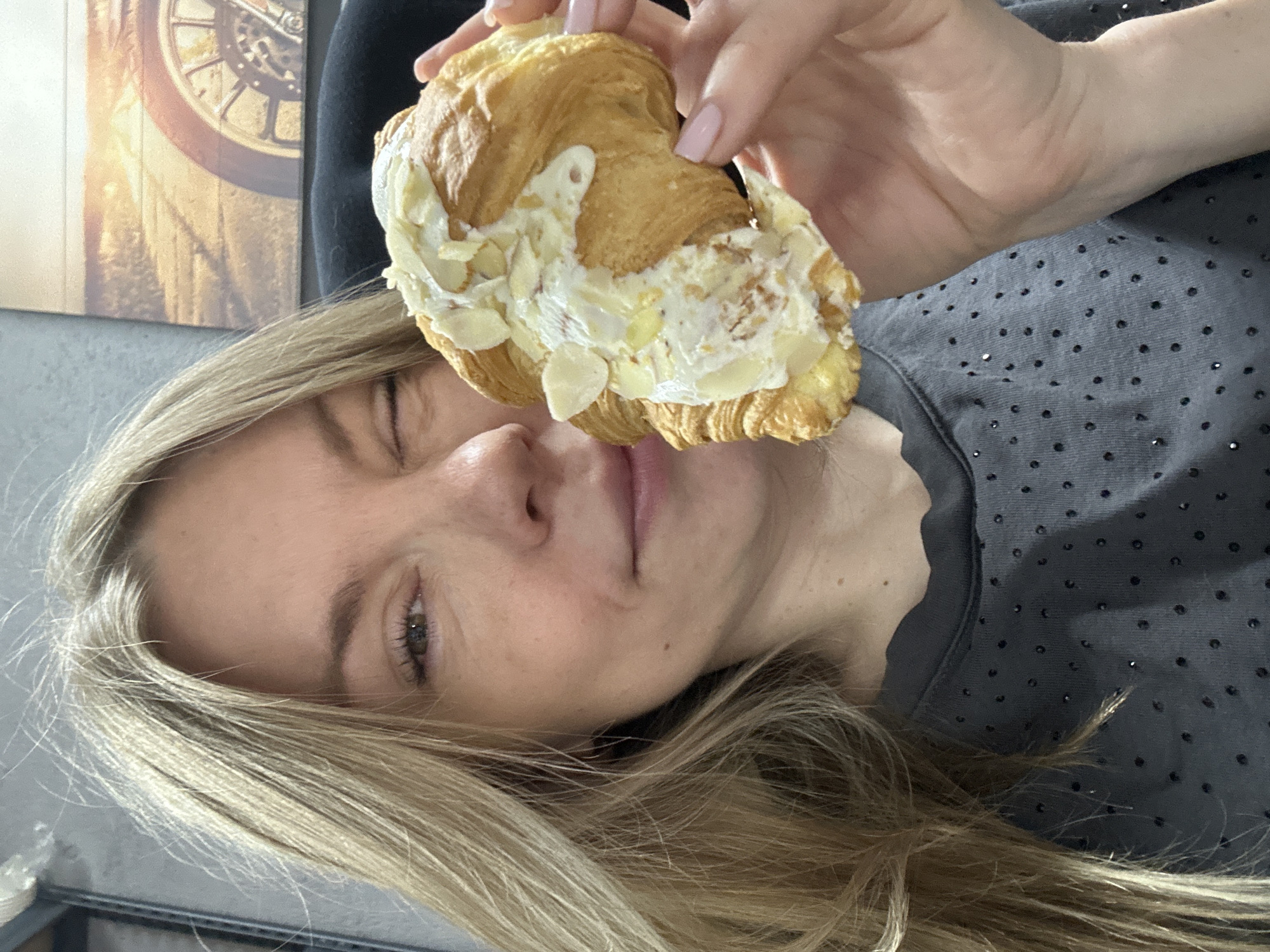 In a close-up, waist-up shot, a young woman with blonde hair is holding a cream-filled croissant to her face, with her left eye winking. She is wearing a dark grey t-shirt adorned with small, scattered rhinestones. The croissant appears to be generously topped with cream and slivered almonds. Behind her, a framed picture hangs on the wall, depicting a close-up of a motorcycle wheel and a road with a sunset in the background. The environment suggests an indoor setting, possibly a home or office, with a grey textured wall visible to the left and the dark backrest of a chair to the right. There is no indication of the location being Blagoveshchensk, Russia.
