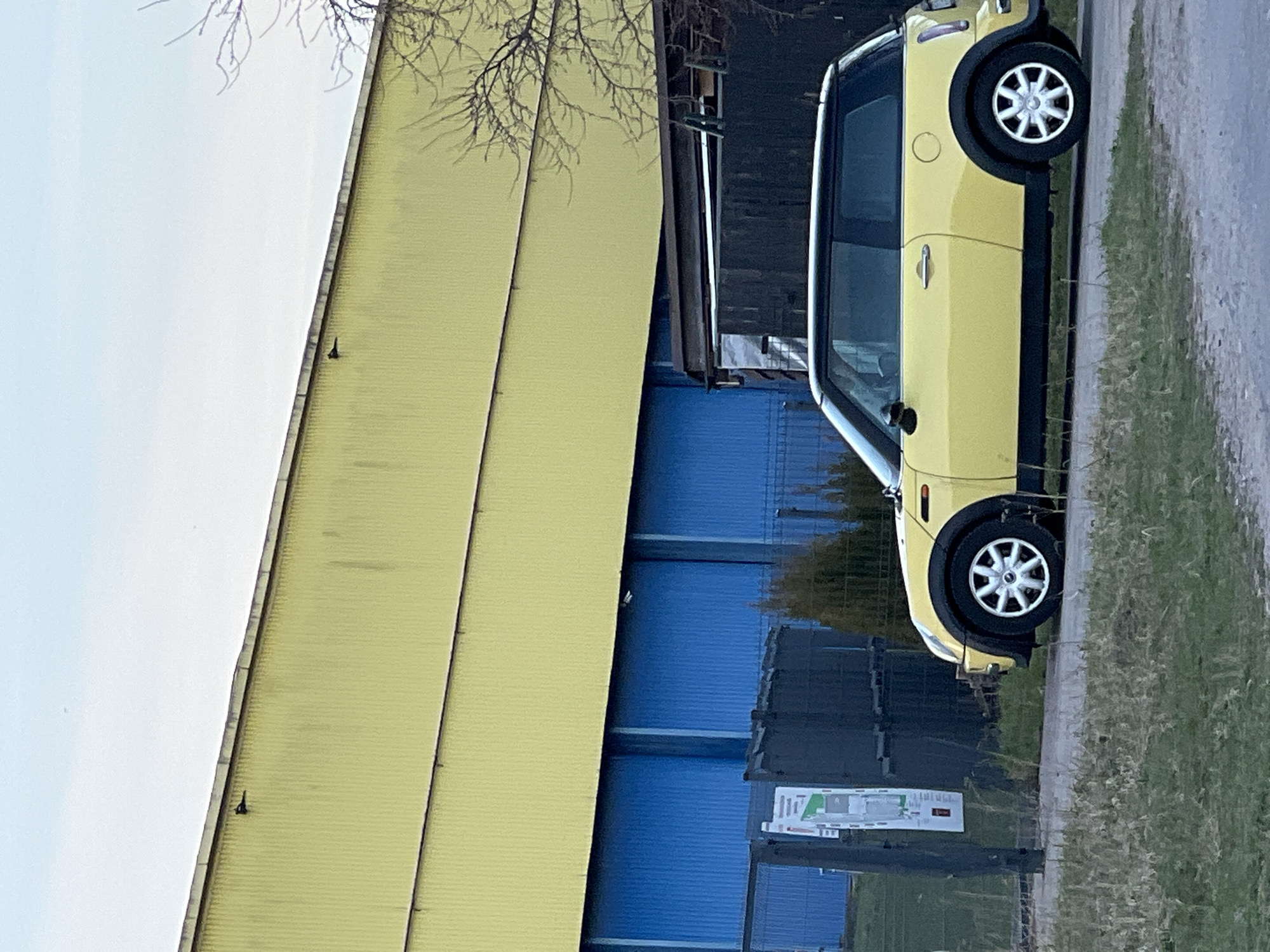 The image depicts a yellow and black Mini Cooper parked on the edge of a grassy area, alongside a building with a yellow corrugated metal exterior. Next to the yellow section of the building, there is a blue corrugated metal section. The context of the city of Riga, Latvia, is not clearly identifiable from the visual information alone, but can be inferred from the location coordinates. There are no people visible in the image. A sign with a map is visible near the base of the building. The sky is visible in the upper-left corner and appears pale. A bare tree branch is visible extending over the yellow building.
