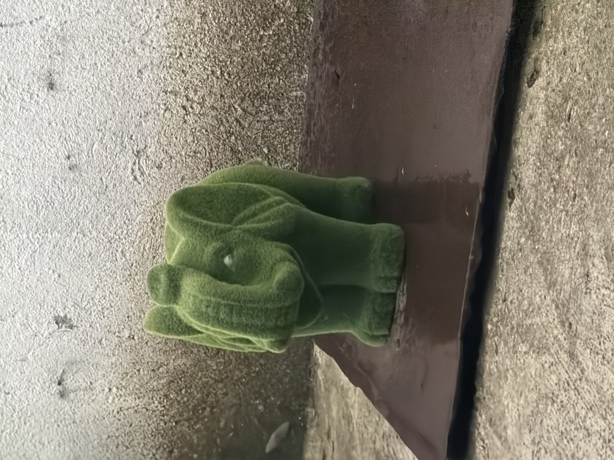 A green, flocked elephant figurine is centrally positioned on a dark brown, rectangular tray with a slightly reflective surface. The figurine faces forward and slightly left, displaying a textured, moss-like appearance, and has a small white mark on its upper trunk. The tray rests on a rough, textured concrete ground, which exhibits varying shades of grey and brown, consistent with weathering and dampness. Behind the tray is an uneven, textured wall; its upper section is light grey with visible imperfections and darker stains, transitioning to a darker grey-brown at the lower section. No individuals are present. The scene is located in Dabene, Bulgaria.
