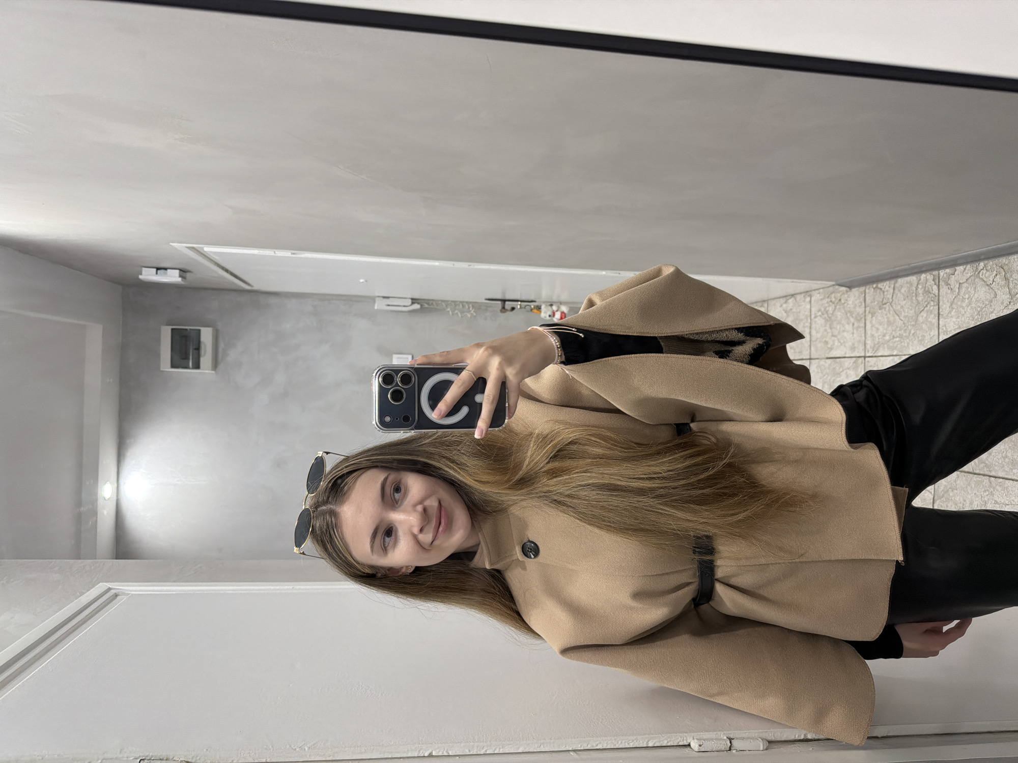 The image depicts a young woman taking a selfie in what appears to be an indoor setting, likely a corridor or hallway. The walls are painted in shades of white and grey, and there's a white door visible. There's also a small, rectangular vent or air conditioning unit on the wall. The woman is wearing a tan coat with a belt, and dark-colored pants or leggings. Sunglasses are perched on her head. She is holding a phone in her right hand to take the picture. Without specific details or context within the image, it's impossible to determine if it's Plovdiv, Bulgaria. No specific landmarks or objects indicate the location.