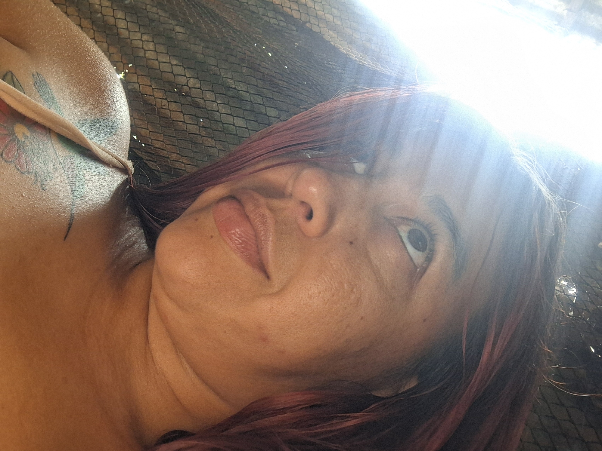 A close-up, high-angle shot captures a woman lying down, looking upwards and slightly to the right. Her head is tilted, giving a left-side profile of her face. She has reddish-brown hair, and her lips are slightly parted. Her left eye is visible, looking up. Her skin appears to be a medium tone, showing natural textures and a few small marks.
On her left shoulder and upper arm, a vibrant tattoo is visible, featuring green and pink/red floral elements, possibly a flower and leaves. She is wearing a light-colored, thin strap, likely part of a top or swimsuit, which runs over her left shoulder.
The background behind her head and upper body consists of a dark, grid-like mesh pattern, possibly a hammock, net, or textured surface. A strong, bright light source, likely the sun, is positioned in the upper right portion of the image, causing significant glare and lens flare that illuminates her hair and the background. This intense light suggests it is daytime and sunny.
No text is visible in the image. The setting, with the strong sunlight and mesh background, could imply an outdoor, relaxed environment, consistent with the provided context of Vallecito, Venezuela.