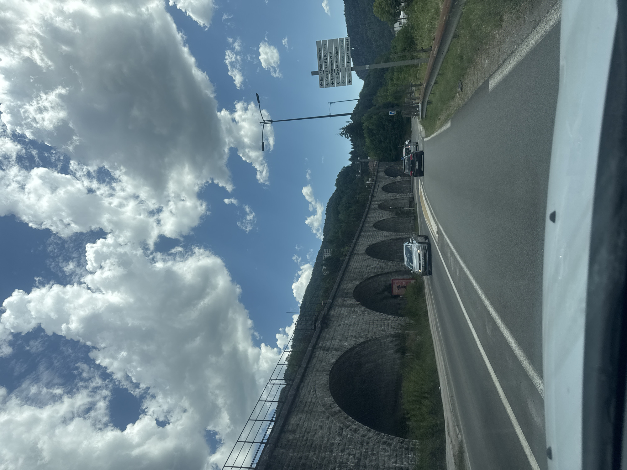 This is a vertical, eye-level, medium shot taken from a vehicle. The scene depicts a road with a viaduct on the left and lush green hills in the background. The sky is bright blue with dramatic white clouds, suggesting a sunny day.
The viaduct is constructed of large, grey stones and features a series of arched openings. A metal fence runs along the top of the viaduct. To the right of the road, there is a green, grassy embankment with some trees. Several cars are visible on the road, suggesting traffic. A tall lamppost with two lights stands near the road.
A prominent signpost on the right side of the road displays several directional arrows and French text, including "Sur Le Puits-Villedieu," "Gare S.N.C.F & Routière," "Gendarmerie PGM," "Crèche Municipale," "La Maison," and "France services." This text provides location context, indicating the presence of services and destinations within the Hauts de Bienne region of France.
The overall impression is a scenic drive through a mountainous or hilly area on a pleasant day. The image captures a moment of travel, with a focus on the infrastructure and natural beauty of the location. The perspective from inside the vehicle adds a sense of movement and personal experience to the scene.
