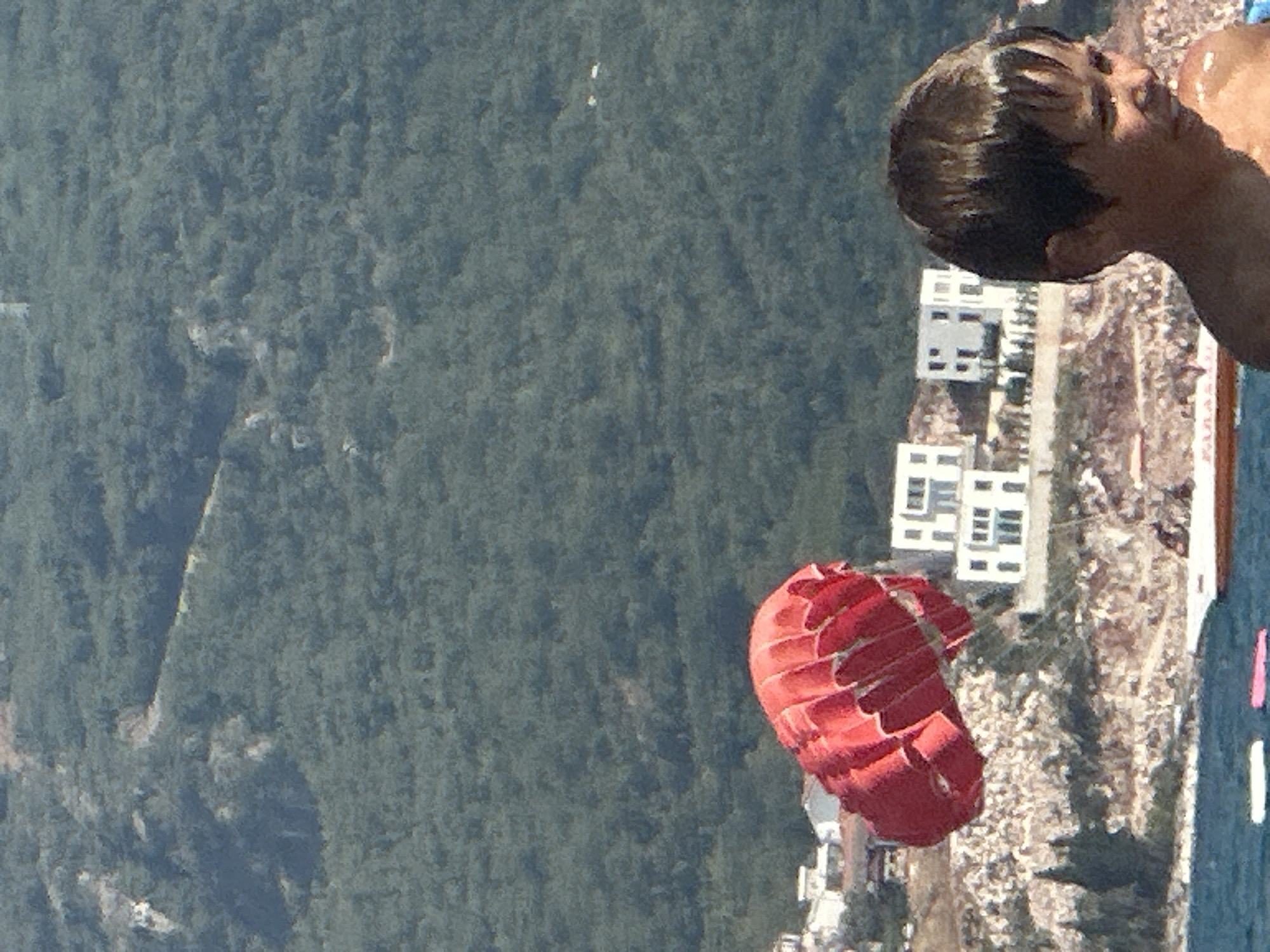 In Boreti, Montenegro, a young boy with short, dark, wet hair looks to his right, his face illuminated by sunlight, as a large, bright red parachute, likely for parasailing, is partially visible in the lower left of the frame. In the background, a lush, green, tree-covered mountain slopes upward, with modern white buildings clustered on a hillside overlooking the water. The water is a deep blue, with a white and red boat visible. The scene captures a moment of summer activity near the coast.