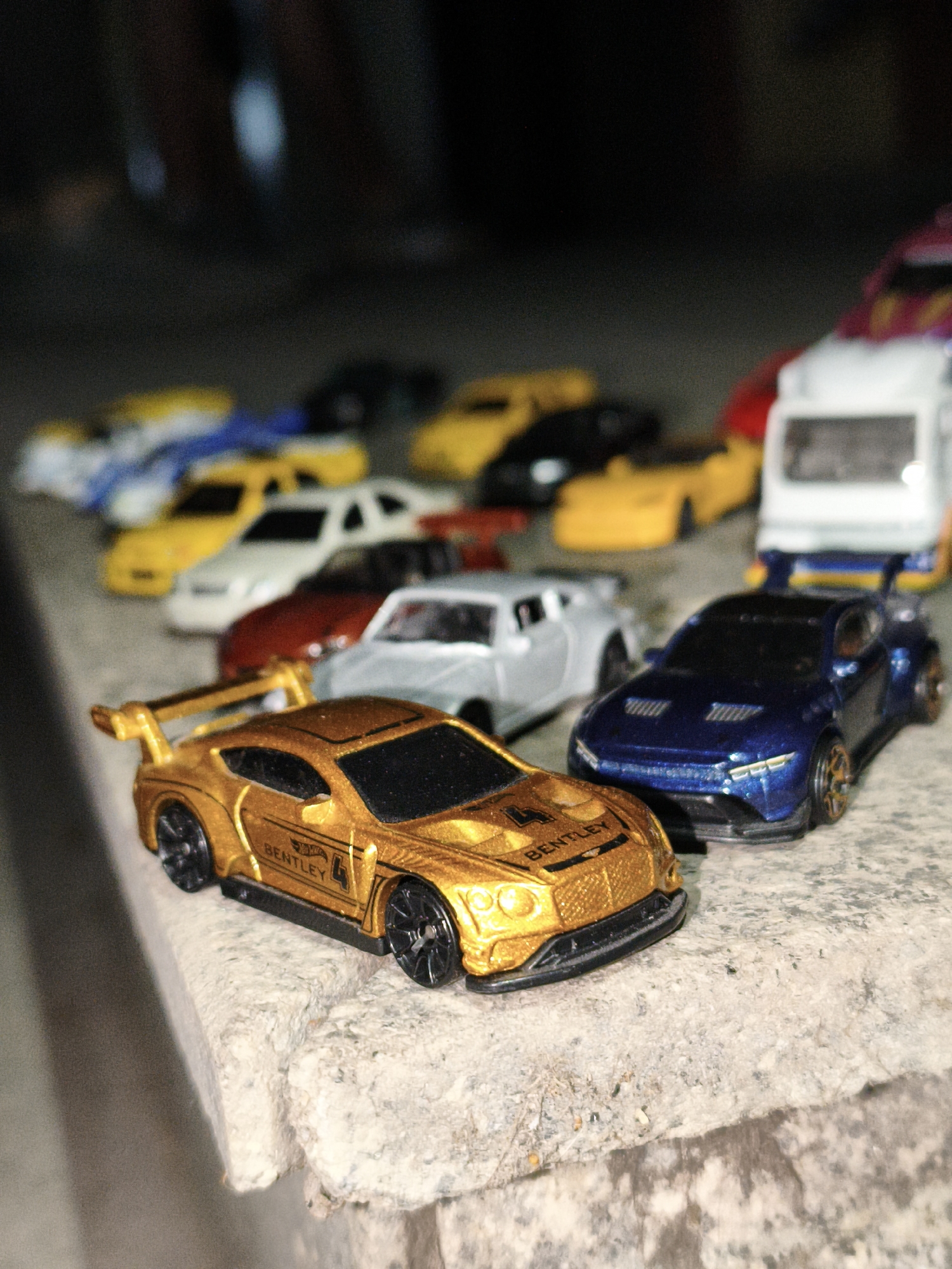 The image shows a collection of toy cars arranged on a stone surface, possibly a ledge or curb. In the foreground, a gold Bentley toy car is positioned next to a blue sports car toy. Behind them, a group of other toy cars is visible, including white, yellow, and silver models, along with what appears to be a toy truck. The background is dark and out of focus.