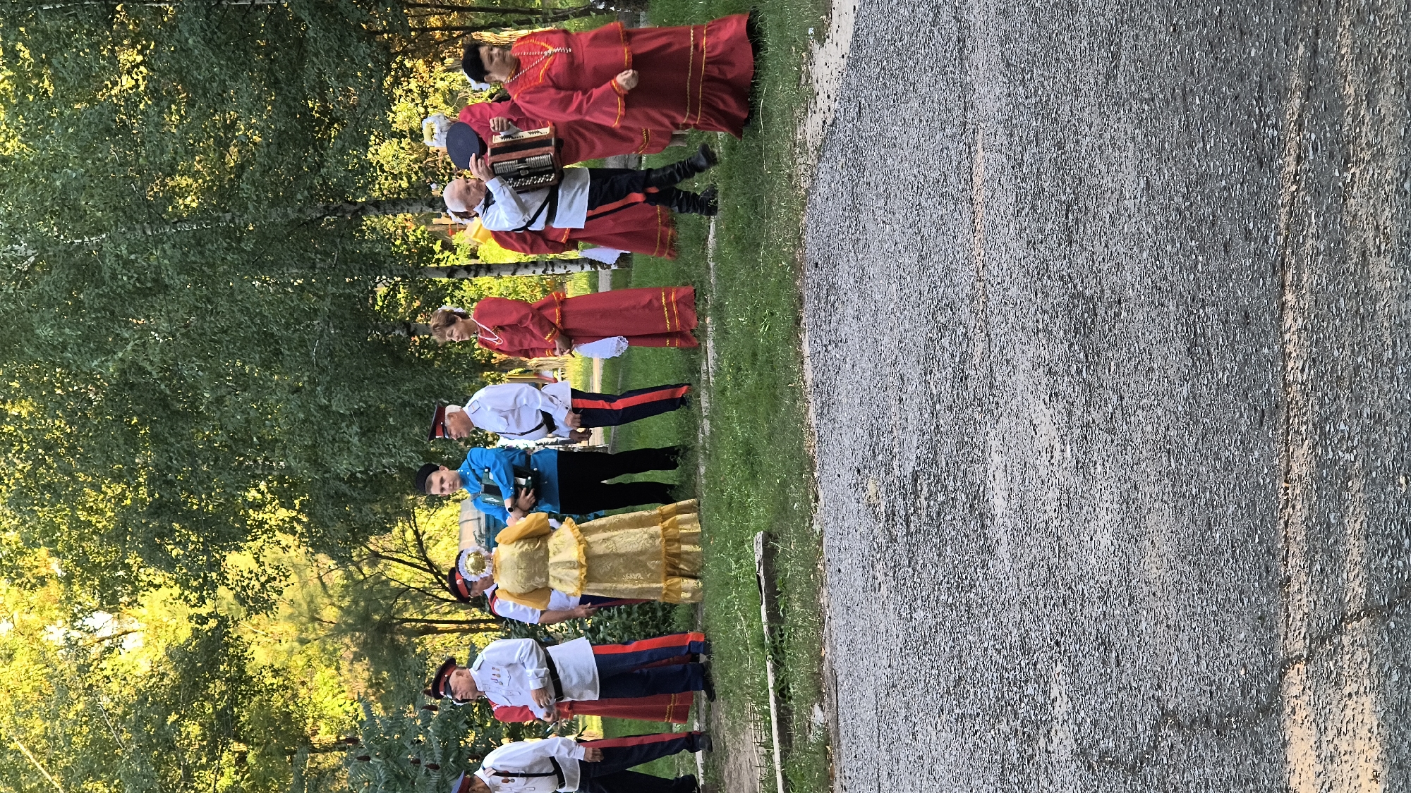 In Mikhaylovka, Russia, a group of people in traditional attire are gathered outdoors in a grassy area with trees in the background. On the left, several men in white uniforms with red and blue trim stand in a line. To their right, a woman in a gold dress is facing them. Next to her is a man in a bright blue uniform. Further to the right, a woman in a red dress stands, and next to her, an older man plays an accordion. Two more women in red dresses are also present, one to the right of the accordion player. The ground in the foreground is a gravel path. The lighting suggests it is daytime, possibly late afternoon, with sunlight filtering through the trees.