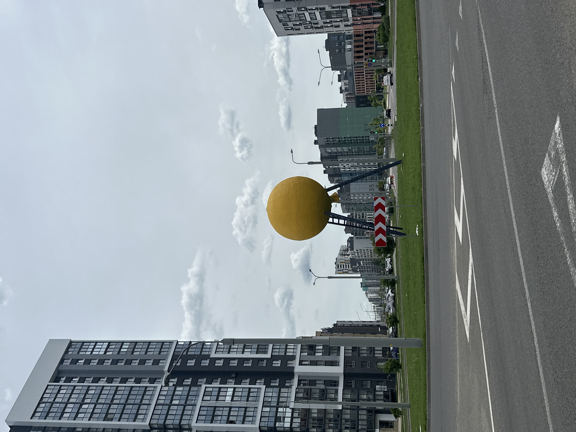 The image shows a street scene in Бараўлянскі сельскі Савет, Belarus. A large, yellow spherical sculpture stands on a support structure in the median of a road. The road has two lanes in each direction, with dashed white lines dividing the lanes. On the left side of the road, a tall, modern apartment building with a dark and light gray facade stands. On the right side, there are more apartment buildings of varying heights and colors, including green and brown. A red and white road sign is visible next to the sculpture. The sky is overcast with clouds. Several streetlights are visible along the road.