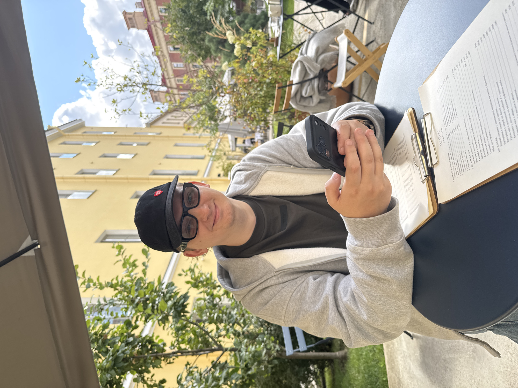A young person wearing a grey hooded jacket, a black baseball cap, and glasses is sitting at a round, dark blue table outdoors, under a beige awning. They are holding a black smartphone in their hands. In front of them on the table are two clipboards with menus. The background shows a courtyard with trees, bushes, chairs, and yellow and red buildings typical of Saint Petersburg, Russia. The sky is visible above with clouds.