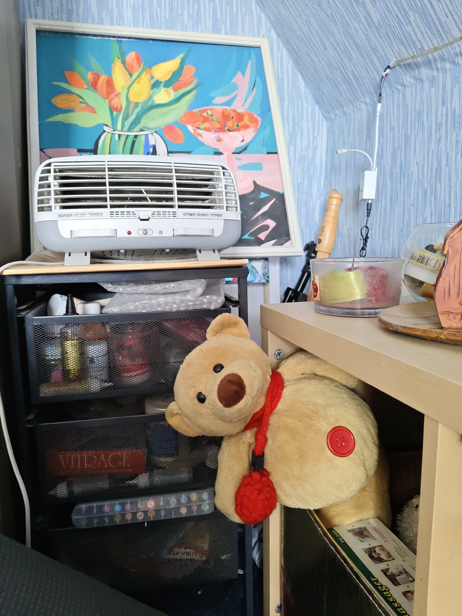 The image depicts an indoor scene with a stuffed teddy bear partially obscuring a wooden shelf. On the shelf, a glass container holds two candles, one yellow and one red, and a small wooden object resembling a pestle. Behind these items, a larger glass bowl contains various objects, some of which appear to be labels or small items. To the left of the shelf, a black metal mesh drawer unit is visible, containing assorted items including bottles, a red rectangular box with text that appears to read "VITRAGE," and a clear plastic case filled with colorful markers. A white electric heater with a large grill is situated on top of the drawer unit. Above the heater hangs a framed abstract painting of tulips in a vase against a blue background. The overall environment suggests a storage or display area. There is no discernible information about the city of Rostov-on-Don, Russia, or any human presence within the image.