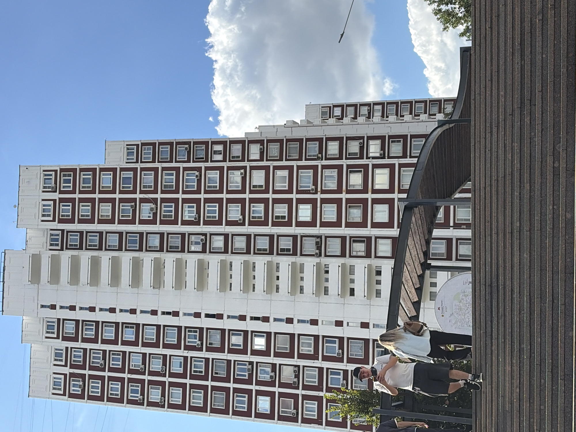 In Moscow, Russia, a multi-story apartment building with a distinctive red and white facade stands against a bright blue sky with scattered clouds. The building is tall and appears to be a residential complex, featuring numerous windows and balconies. In the foreground, a wide set of granite stairs leads up to a covered walkway. Two people are visible on the walkway: a man in a white t-shirt and dark shorts, holding a mobile phone, and a woman with blonde hair wearing a light blue jacket and dark pants, carrying a backpack. Behind them, a large circular map is displayed. The overall scene captures a typical urban environment on a sunny day, with people interacting with their surroundings in front of a prominent architectural structure.