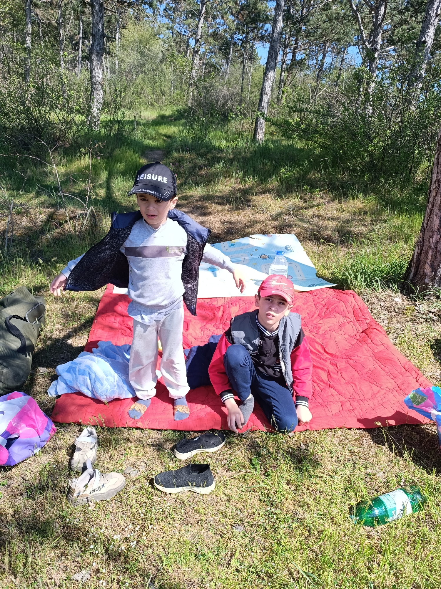 Two boys are positioned on a red blanket spread out in a grassy wooded area. The boy on the left is standing, wearing a grey shirt, white pants, slippers, and a black cap with the word "LEISURE" on it. He's in the process of putting on a black jacket. The boy on the right is kneeling, wearing a red cap, a black jacket, a red vest, and blue pants. Beside the blanket are a few items: shoes, a bag, a blue blanket, and a bottle. A white and blue mat with a bottle on it can be seen behind the standing boy. The area is wooded with sunlight filtering through the trees.