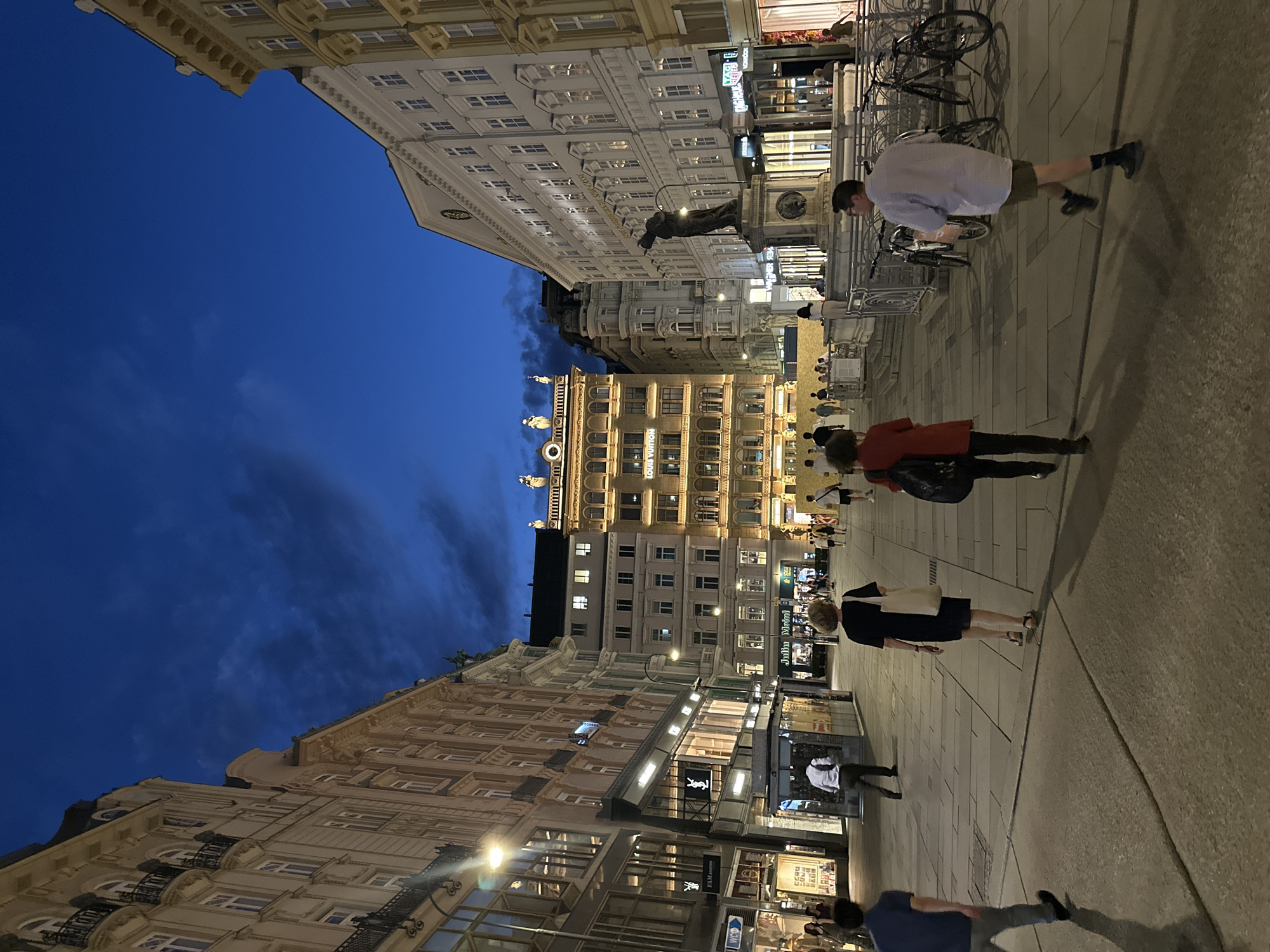 This image captures a street scene in Vienna, Austria, at dusk. The architecture is characteristic of the city, with grand, ornate buildings lining a wide pedestrian plaza. The sky is a deep, dark blue with scattered clouds, indicating twilight. The buildings are illuminated, highlighting their classical facades, balconies, and windows. Storefronts are visible along the street level, with some illuminated signs, including one for Louis Vuitton. Several people are walking in different directions across the plaza. In the foreground, on the right, a man in a light blue shirt and khaki shorts is walking away from the camera. To his left, a woman in a dark dress and a light-colored tote bag is also walking away. Further left, a person in a dark blue shirt and grey pants is walking towards the left. In the middle ground, there are more people walking, and a statue on a pedestal stands prominently in front of a large white building. Bicycles are parked near the statue. The ground is paved with large, light-colored stone slabs. The overall impression is of a lively urban environment transitioning from day to night, with historical architecture providing a rich backdrop.