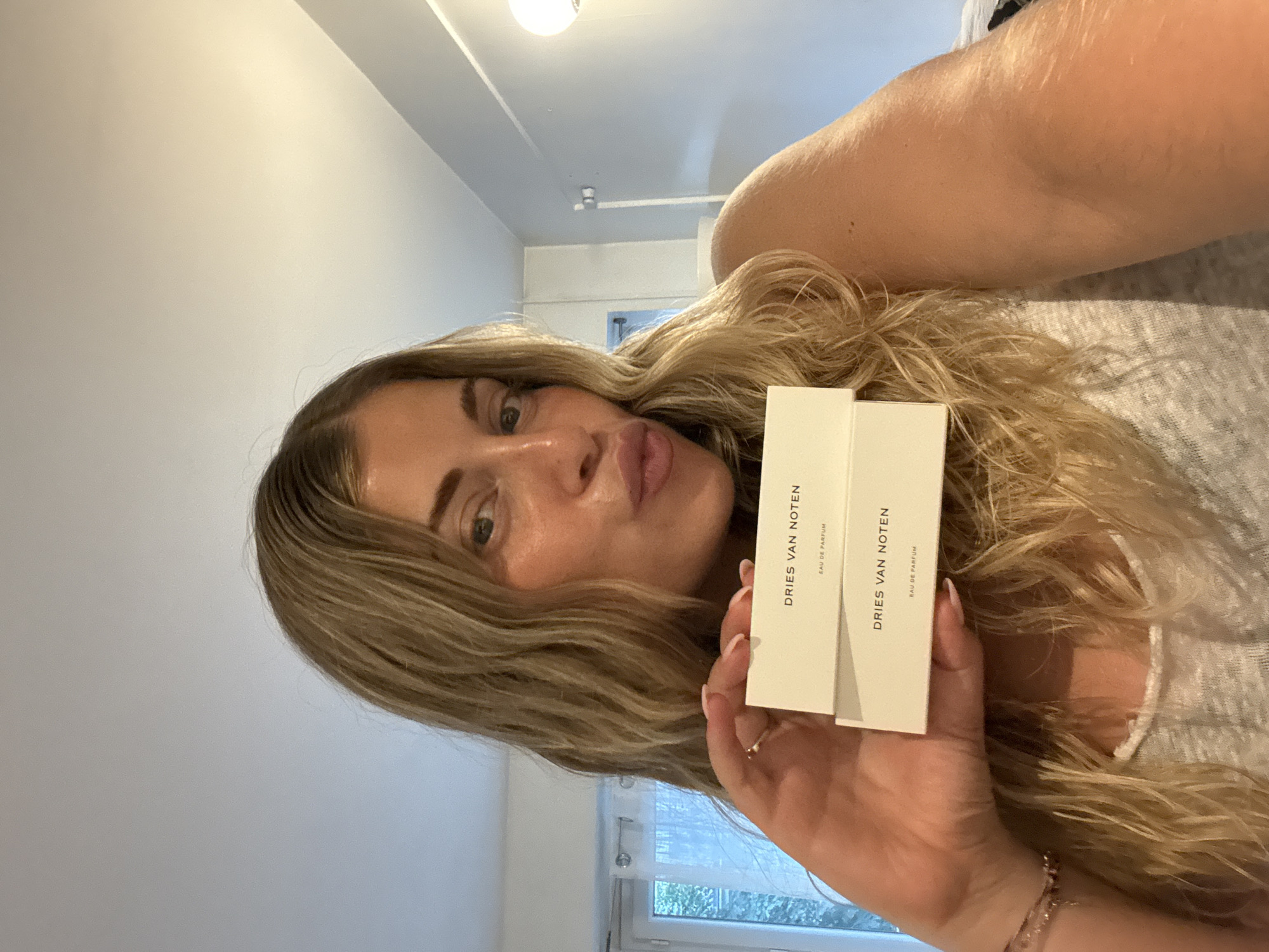 A woman with long blonde hair is holding two white rectangular boxes with the text "DRIES VAN NOTEN EAU DE PARFUM" printed on them. The woman is looking at the camera with a slight pucker of her lips. She is wearing a grey top and a bracelet on her right wrist. The background includes a white ceiling with a light fixture, a light grey wall, and a window with a view of green foliage. The location appears to be indoors and is not identifiable as Paris, France from the visual information provided.