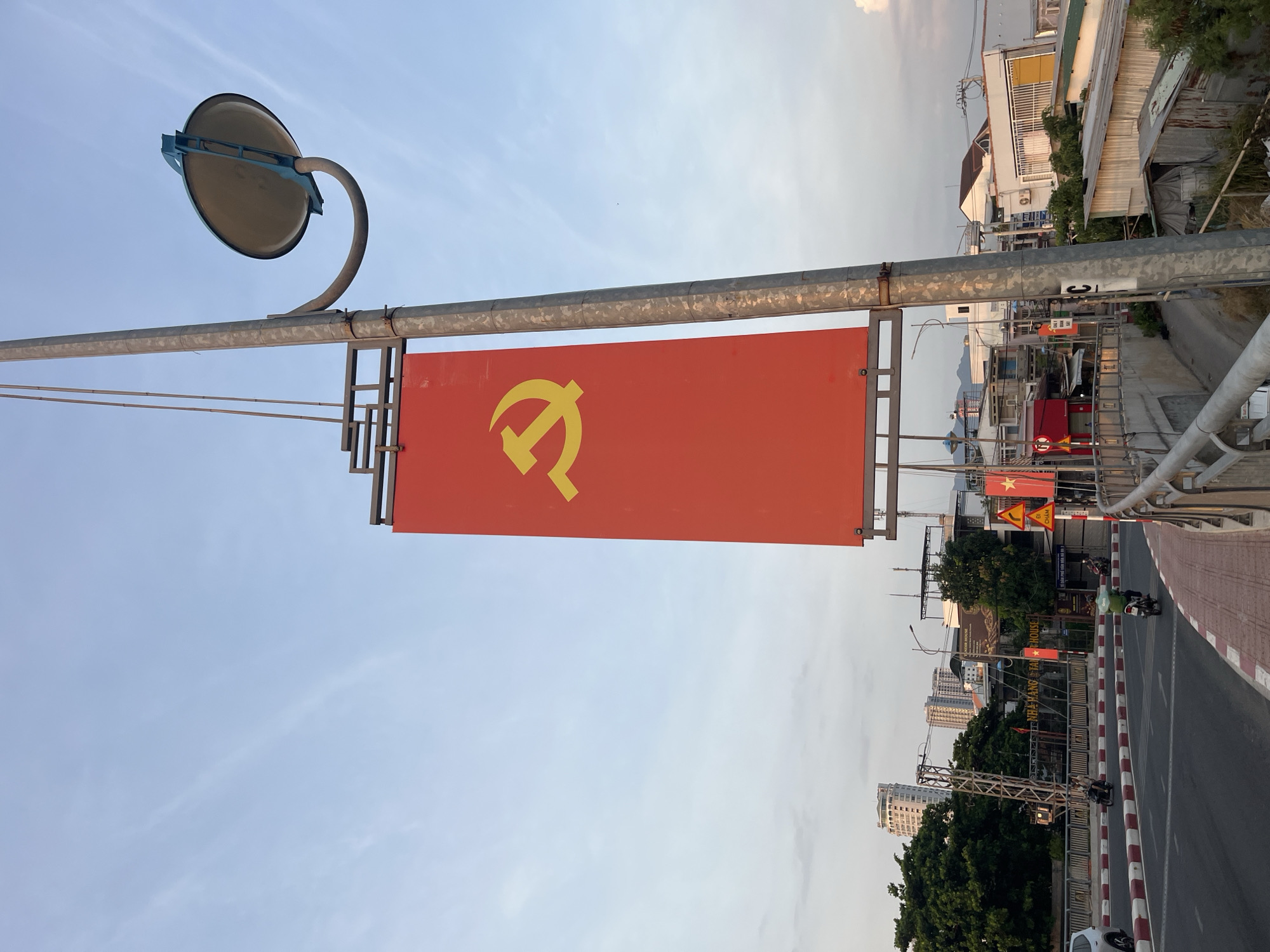 The image shows a red banner with a yellow hammer and sickle symbol hanging from a metal pole in Bac Nha Trang Ward, Vietnam. The banner is rectangular and prominently displayed. A circular mirror is attached to the pole above the banner. In the background, there's a road with red and white markings, buildings of varying heights and conditions, including some with corrugated iron roofs, and trees. Several Vietnamese flags are also visible along the road. The sky is light blue and slightly overcast. A motorcycle can be seen on the road.