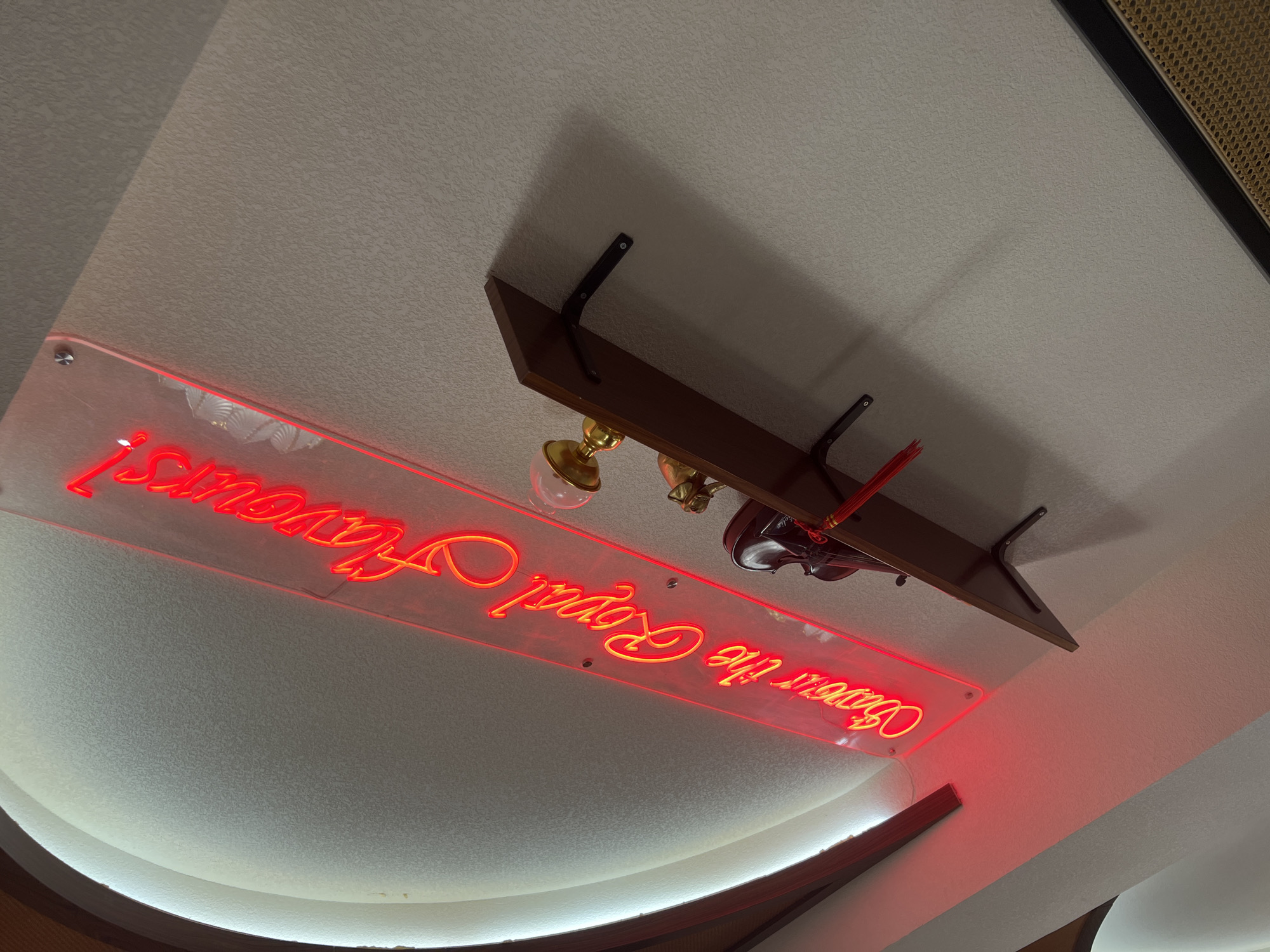 An upward-angled, tilted photograph captures an interior wall in the Chongqing High-tech Zone, China, featuring a red neon sign and a mounted shelf. The central element is a red neon sign, illuminated brightly, reading "Savour the Royal Flavours!" in a flowing script, mounted on a clear acrylic panel. The sign is affixed to a light-colored, textured wall. Below the sign, a dark wooden shelf is horizontally mounted to the wall with two visible black metal brackets. Objects arranged on the shelf include a golden, ornate lamp with a spherical top, and a dark, possibly red-brown, decorative box or container adorned with a red tassel. To the far left, a curved architectural element, possibly a ceiling cove or recessed archway, is illuminated from within, creating a bright white glow along its edge and bordered by dark wood trim. The wall exhibits a consistent stippled or speckled texture. A shadow from the shelf is cast on the wall below it. In the bottom right corner, a section of a woven, possibly rattan or wicker, panel with a dark frame is partially visible. No people are present.