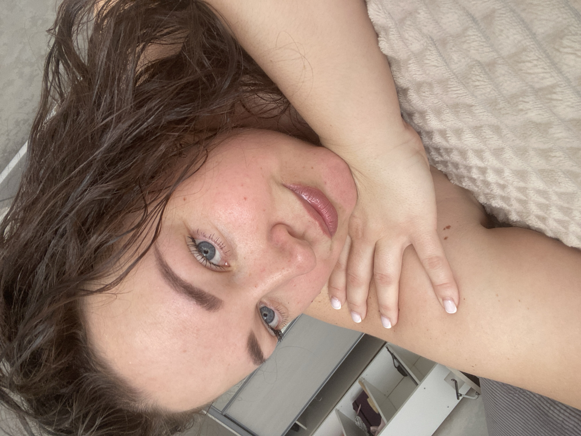 A woman with wet, dark hair and blue eyes is lying down. Her right hand is near her neck. The background includes a light-colored, textured surface and part of a white cabinet or shelving unit. The location is not discernible from the image.