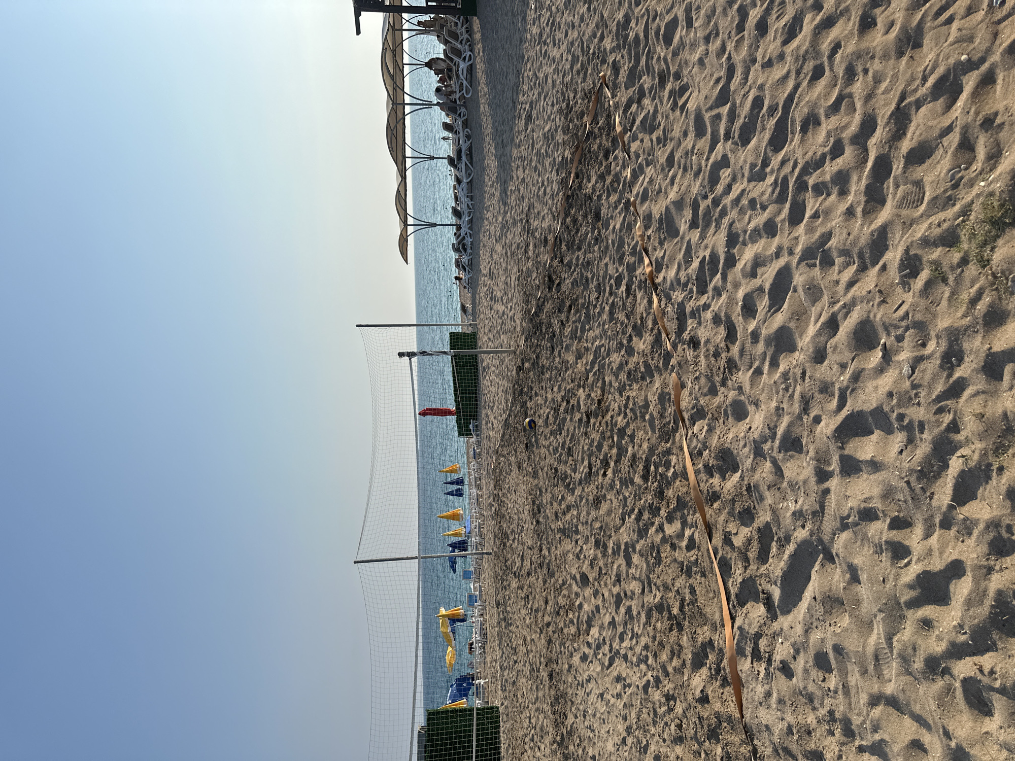 This is a medium-shot, eye-level photograph of a sandy beach in Manavgat, Turkey, under a clear blue sky. In the foreground, a volleyball net stretches across the frame, supported by poles. A volleyball rests on the sand to the right of the net. A brown ribbon or cord is laid out on the sand, forming a wavy line.
In the midground, beyond the net, the blue ocean meets the horizon. Along the shoreline, rows of white lounge chairs are arranged under large, canvas canopies, providing shade. Several beach umbrellas, in yellow and blue, are also visible. A few people are seated on the lounge chairs, and some are in the water.
The environment is characteristic of a resort beach, with amenities for relaxation and recreation. The sand is fine-grained, and the overall impression is one of a sunny, peaceful day at the seaside.