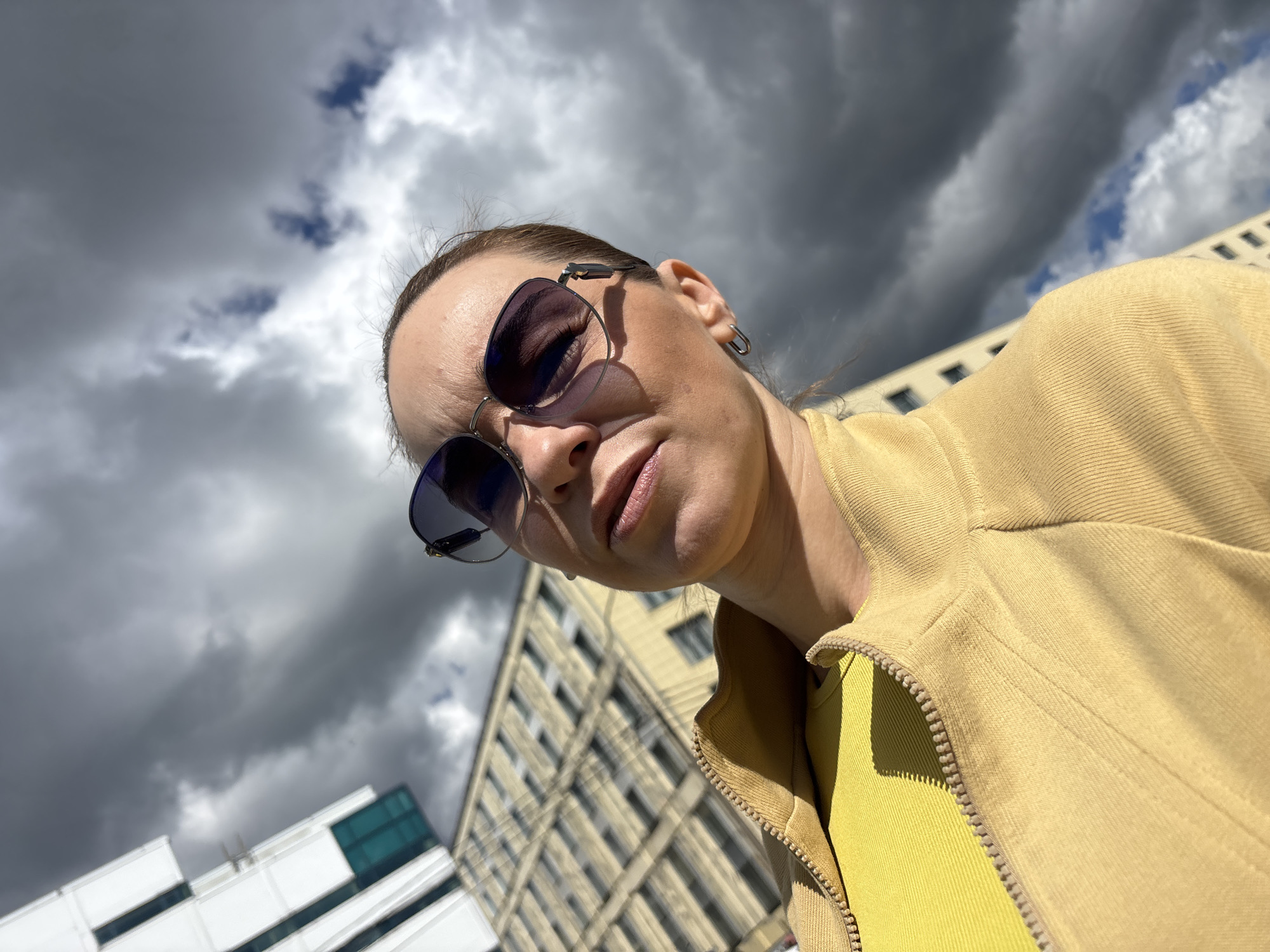 A low-angle, close-up, outdoor selfie captures a woman in Saint Petersburg, Russia, against a backdrop of a building and a dramatic, cloudy sky. The woman, wearing sunglasses and a yellow top underneath a tan zippered jacket, looks directly at the camera with a neutral expression. Her hair is pulled back, and she has hoop earrings. The building behind her appears to be a multi-story structure with a beige facade and dark windows. The sky is filled with dark, heavy clouds interspersed with patches of blue sky. The image is sharp and well-lit, with the sun casting shadows.