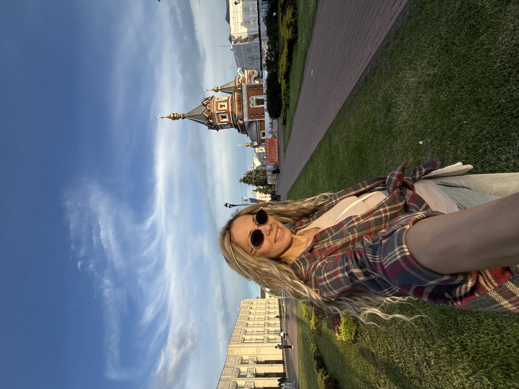 The image, rotated 90 degrees clockwise, captures a woman taking a selfie outdoors with a distinctive architectural building in the background under a bright sky.
The main subject is a smiling woman, positioned on the right side of the frame (if rotated upright). She has long, wavy blonde hair, wears dark, round sunglasses, and is dressed in a light-colored top covered by a red, blue, and cream plaid flannel shirt tied at her waist. She appears to be wearing light-colored jeans or trousers. Her expression is cheerful and relaxed.
The setting is a well-maintained urban park or public square. In the mid-ground, to the left (if upright), stands a prominent red-brick building with intricate white trim, arched windows, and multiple dark-colored domes crowned with golden spires. This architecture strongly resembles a Russian Orthodox church or cathedral. Behind this structure, another building is partially visible, adorned with a banner displaying Cyrillic text and a graphic that appears to be a St. George ribbon, suggesting a historical or patriotic context. Further in the background, a large, light-colored, multi-story building with numerous windows indicates a city environment.
The foreground (bottom of the image if upright) shows neatly manicured green grass and a paved pathway that curves through the scene. The sky above is a vibrant blue with scattered wispy white clouds. The lighting suggests it is bright daylight, possibly late afternoon or early evening. A red sign with illegible Cyrillic text is visible near the church.