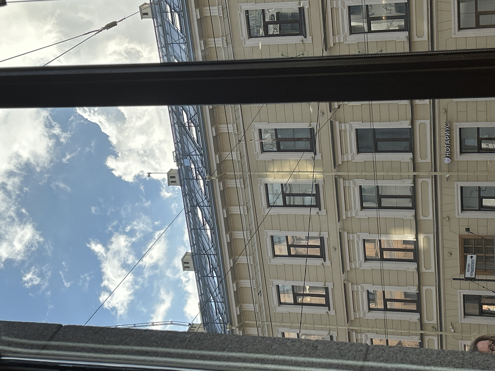This is an eye-level medium shot of a building facade seen through a window with a dark vertical frame in the foreground. The building is light yellow with white decorative trim around the windows and along the roofline. The roof is a dark, sloped structure with visible skylights and structural elements. The sky above is blue with scattered white clouds. Electrical wires are visible crossing the frame in front of the building and the sky. On the building, there are multiple windows, many of which reflect the sky and surrounding environment. Some windows have signage visible inside, and one prominent sign on the lower floor reads "НОТАРИУС" with a small emblem. A street sign with Cyrillic text is also visible near a window on the lower floor. A small security camera is mounted on the facade above the "НОТАРИУС" sign. On the left side of the image, a portion of a grey, textured architectural element, possibly a pillar or wall, is visible. A person's face, partially visible at the bottom left, is looking towards the left, wearing glasses.