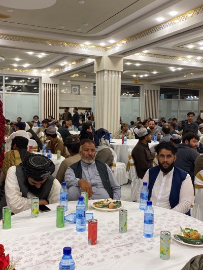 The image shows a group of men seated at tables in a large, decorated room. The location is identified as Kandahar, Afghanistan. The men are dressed in traditional clothing, including turbans and vests. The tables are covered with white tablecloths and set with bottles of water, cans of soda, and plates of food. The room is brightly lit with ornate ceilings and pillars. The overall atmosphere suggests a formal gathering or event.