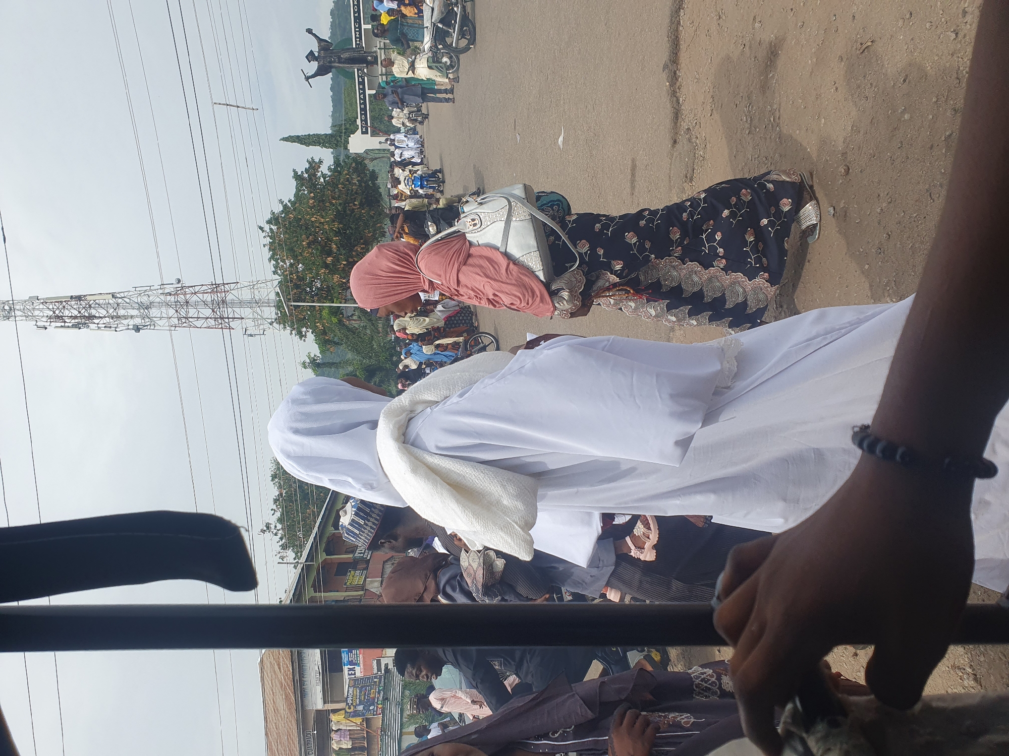 The image depicts a street scene in Lokoja, Nigeria. From an elevated viewpoint, possibly inside a vehicle, the scene shows a crowd of people on a paved street. Individuals are dressed in a variety of clothing styles including traditional garments such as hijab and white robes. A woman wearing a pink headscarf and a black patterned dress stands with a silver purse. Another figure wears a white robe. In the background, a tall communication tower is visible, along with overhead power lines and a monument that shows a graduation statue on a sign that reads "KOGI STATE POLYTECHNIC, LOKOJA". The environment includes trees and buildings along the road. The overall impression is a busy public space in Lokoja.