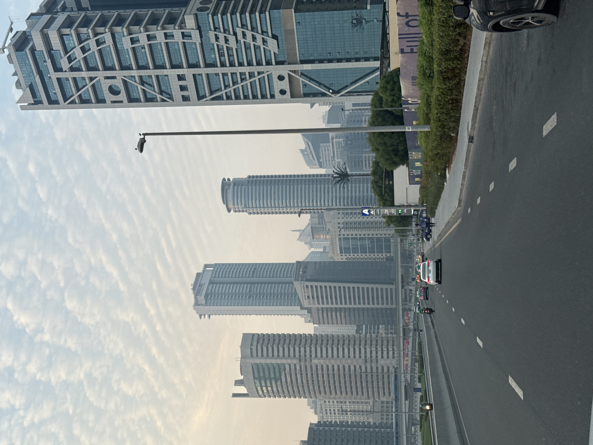 The image captures a view of a multi-lane highway in Dubai, United Arab Emirates, with a cityscape dominated by modern skyscrapers in the background. The sky is overcast with scattered, wave-like clouds. On the right side of the road, a black SUV is partially visible. The highway is marked with white dashed lines. Traffic consists of several cars and at least two motorcycles. On the left side of the highway, there is a concrete barrier and a grassy median. Further left, elevated tracks for a train or metro system are visible, with a train passing on them. Along the right side of the road, there is a dense line of green bushes and trees, with a construction hoarding in the background displaying the text "Full of". Tall buildings, characteristic of Dubai's architecture, are prominently featured, including a distinctive building with a geometric facade on the far right. A lamppost with two birds perched on it is also visible. The overall atmosphere suggests an urban environment during daylight, possibly early morning or late afternoon due to the soft lighting.