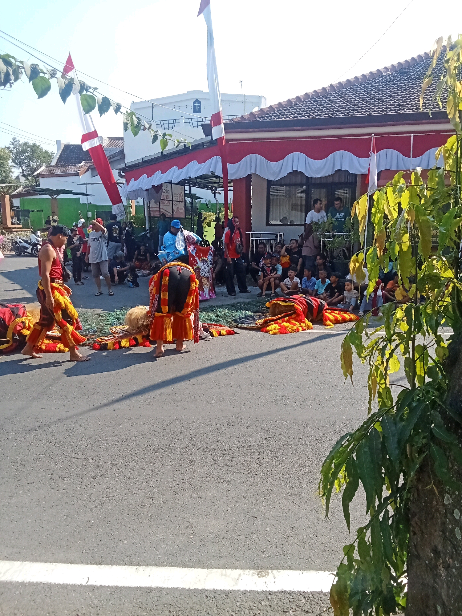 In Magetan, Indonesia, a vibrant cultural procession is unfolding on a paved street under a bright, sunny sky. Two individuals, adorned in traditional, colorful attire of red, yellow, and black, are the focal point. One person, with bare feet, wears a sleeveless red top and patterned pants, while the other is covered head-to-toe in a striking, lion-like costume, also in red, yellow, and black. Both appear to be either preparing for or participating in a performance, with a large, elaborately decorated costume piece lying on the ground between them.
The surrounding environment is bustling with onlookers. Spectators, including men, women, and children, are gathered on the side of the road, many seated on the ground, observing the event. A building with a red and white facade, decorated with festive banners and flags, serves as a backdrop. The Indonesian flag, red and white, is prominently displayed on poles and as part of the building's decorations, suggesting a national celebration or event. Trees with lush green foliage line the right side of the frame, adding a natural element to the urban scene. A motorcycle is parked on the left, indicating the presence of transportation in the area. The overall atmosphere suggests a lively community gathering and cultural expression.