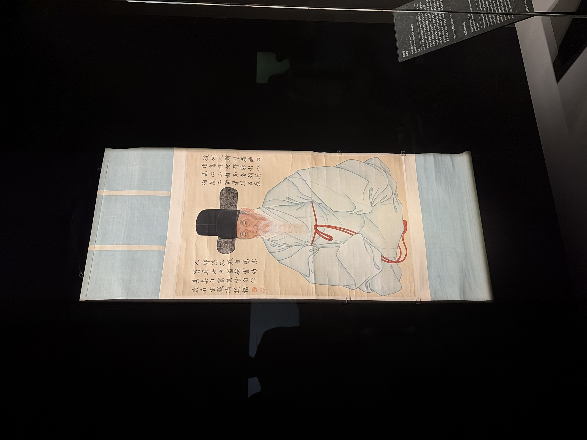 The image shows a vertical scroll painting of a man in traditional Korean attire, displayed in a dimly lit exhibition space. The painting depicts a seated elderly man with a long white beard, wearing a black hat and a pale blue robe tied with a red cord. Calligraphy in Korean characters is present on the right side of the painting, and a red seal is visible on the lower left. The scroll is hung against a black background, with sections of pale blue fabric at the top and bottom, possibly part of the exhibition display. To the right of the scroll, a dark information plaque with text in Korean and English is partially visible. The lighting is subdued, highlighting the painting and the plaque. The overall impression is of an art exhibition setting, likely in a museum. The plaque suggests the artwork is a self-portrait of Kang Se-hwang (1713-1791). The location is specified as Seoul, South Korea, likely within the National Museum of Korea, given the style of exhibition and the presence of Korean historical art.
