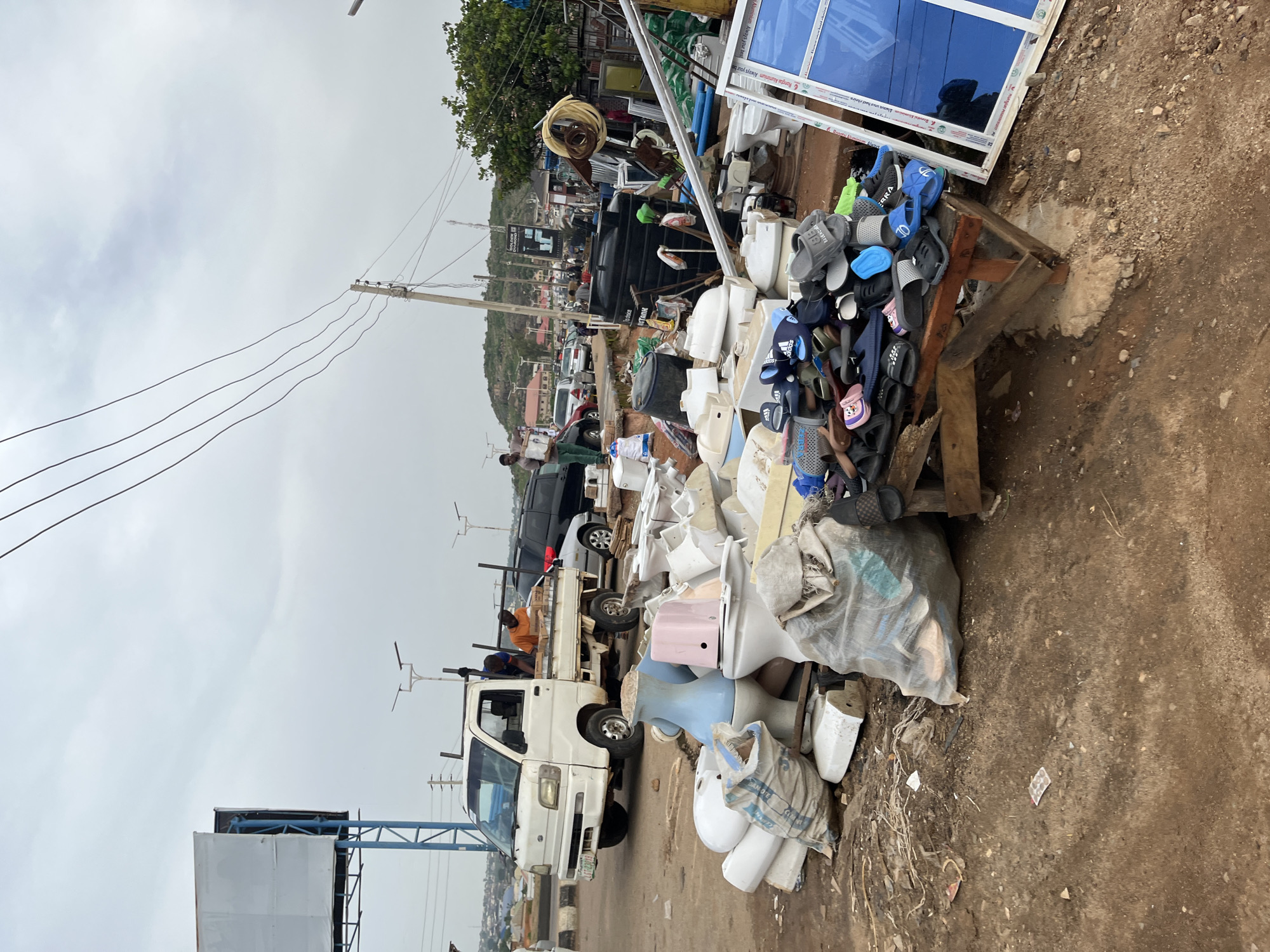 This image depicts an outdoor scene in Gura Top, Nigeria, likely a market or a street vendor area. The time of day appears to be daytime, with an overcast sky suggesting cloudy weather.
The foreground is dominated by a large pile of assorted items, including what appear to be plumbing fixtures like toilets and sinks, as well as a significant collection of footwear, primarily flip-flops and sandals. Some of these items are neatly stacked, while others are in disarray, suggesting either a display for sale or a collection of discarded goods. A large black water tank is visible in the middle ground, as are several vehicles, including a white pickup truck with people in it. The truck is loaded with items, and two individuals are visible in the back.
In the background, there are buildings and a hillside densely populated with structures, indicating an urban or semi-urban environment. Electrical wires are prominent in the sky, strung between utility poles, a common sight in many Nigerian towns. The ground is a mix of dirt and paved surfaces. There is visible text on a sign in the background which reads "GOLDEN DIAMOND". Another sign with blue accents is partially visible on the left side of the image.
The overall impression is one of a busy, functional area where goods are being sold or processed. The presence of both new-looking items and potentially discarded ones could suggest a variety of activities, from a plumbing supply store to a recycling or second-hand goods operation. The scene is bustling with activity, implied by the presence of vehicles and people.