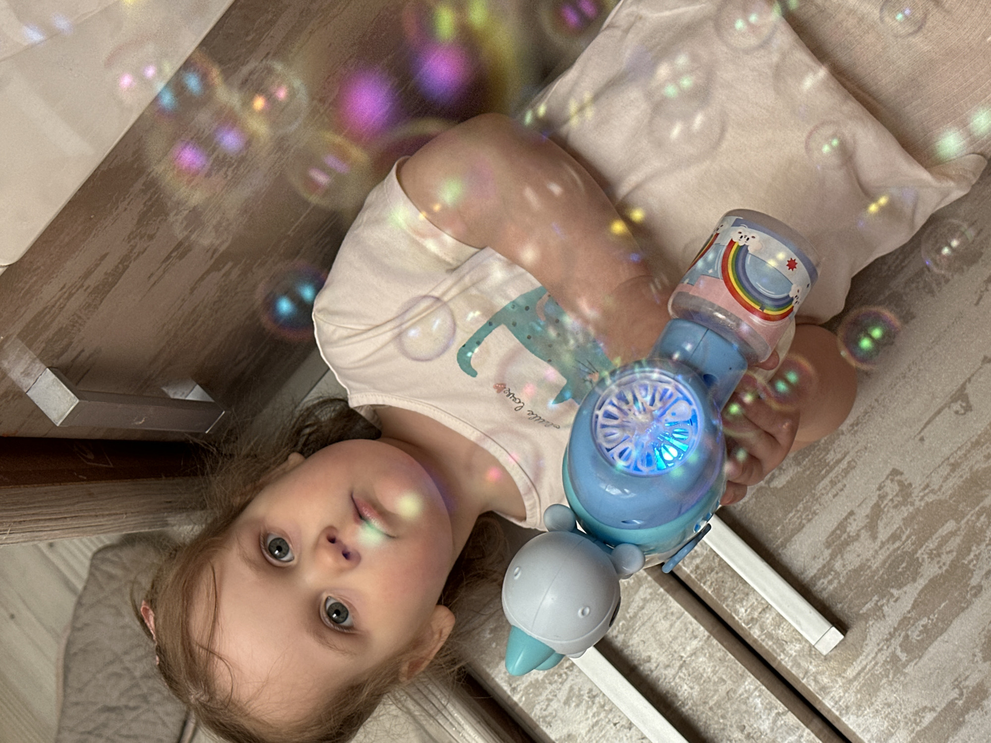 A young child lies on their back on what appears to be a wooden floor or surface indoors, near the leg of a table or other furniture. The child holds a bubble-blowing machine in their right hand. Numerous iridescent bubbles are visible in the air, suggesting the machine is in operation. The location is the city of Chaikovsky, Russia.