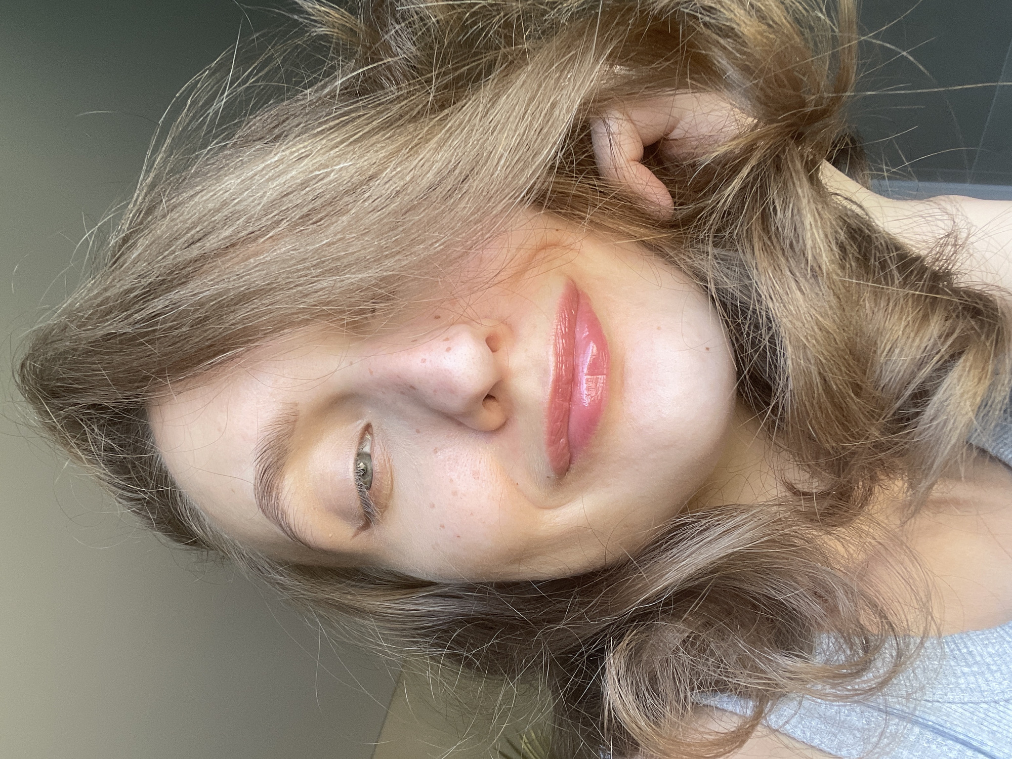 A close-up, angled portrait shows a woman with light brown and blonde hair. Her skin is fair, with freckles visible on her nose and cheek. She has a green-gray eye that is visible, and her lips are coated in glossy pink lipstick. Part of her hand is visible near her ear, suggesting she is touching her hair. The background is a plain, light-colored wall. There is no indication of the location being in Подшивалово, Russia.