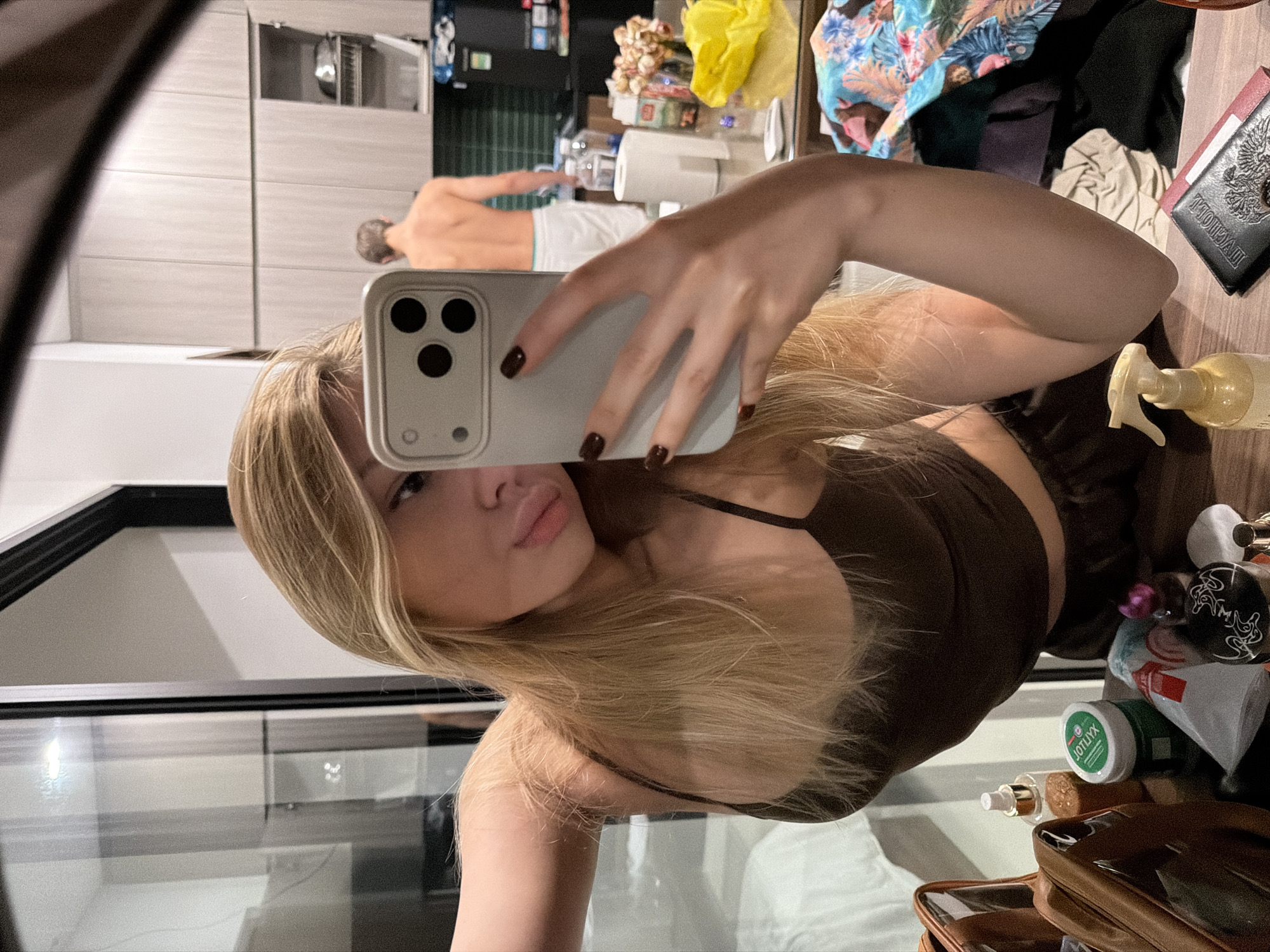 A young blonde woman is taking a selfie in a mirror, her arm holding up a phone. She is wearing a dark brown tank top and matching shorts. In the background, reflected in the mirror, a man with short brown hair and fair skin is standing with his back to the camera, wearing white shorts. The environment appears to be a modern apartment or hotel room with a kitchen area and a large glass door or window. On the surface in front of the woman are various cosmetic items and a passport. The scene suggests a casual, personal moment. The presence of a passport implies international travel. Given the provided location information, this scene is taking place in Đà Nẵng, Vietnam.
