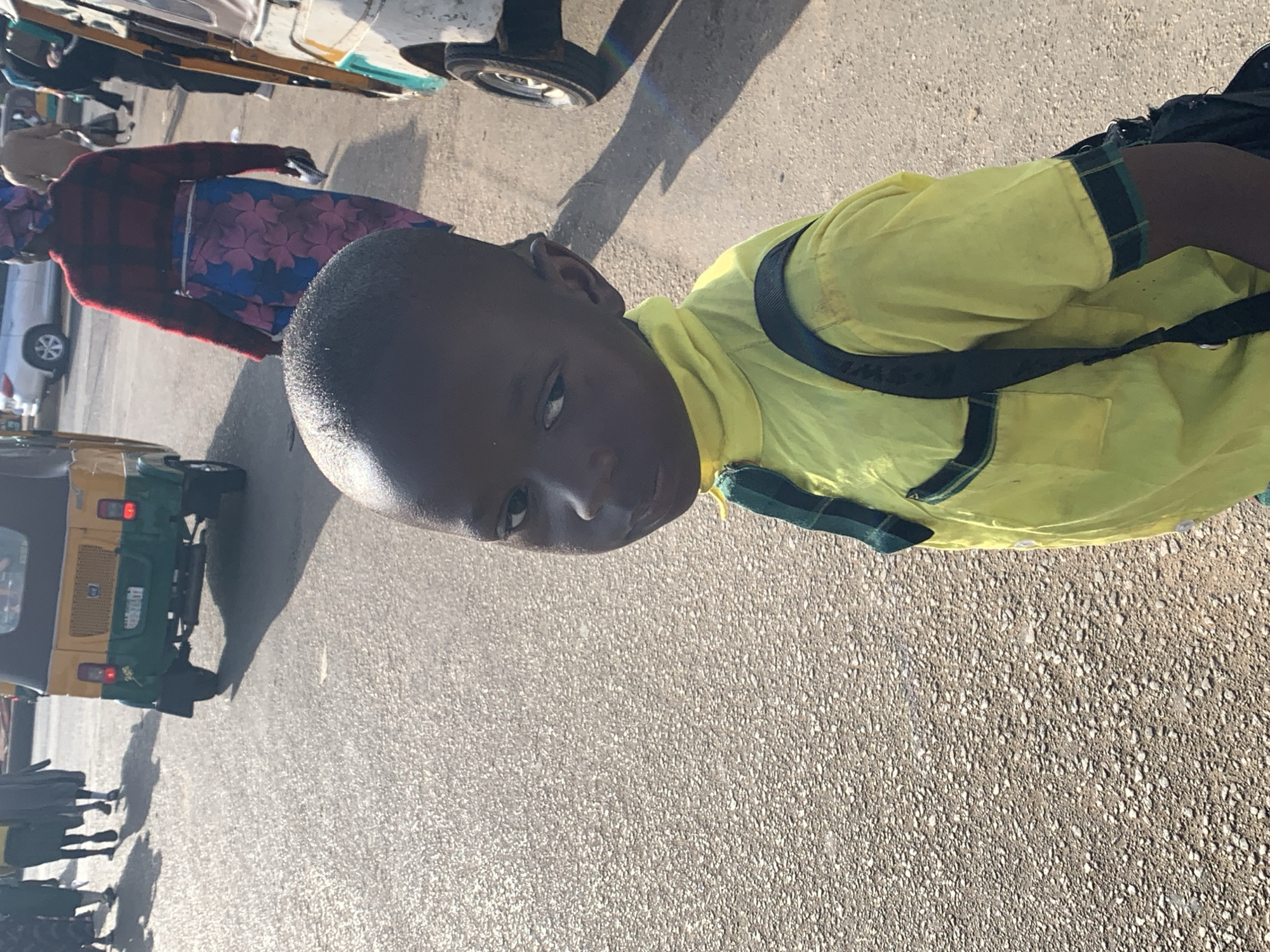 A young boy with dark skin and a backpack stands in the foreground, facing the camera slightly. He is wearing a yellow shirt with dark green and black trim. In the background, other people and vehicles are visible on a paved road. To the left, an auto-rickshaw with a yellow and green body is parked. Further back, a white car is driving on the road. To the right, another auto-rickshaw with a white and yellow body is partially visible. The overall scene suggests a street in a populated urban area, likely in Gura Top, Nigeria, with a busy atmosphere.