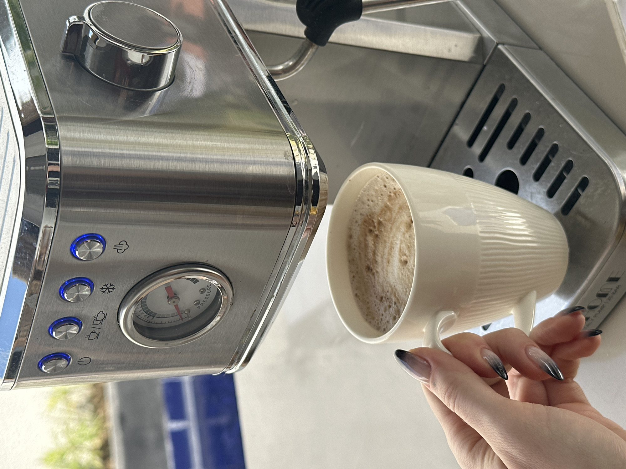 A close-up shot shows a person's hand holding a white ceramic mug filled with frothed coffee, positioned beneath a stainless steel espresso machine. The machine has several illuminated blue buttons and a pressure gauge. The background is blurred, but hints of blue tile and greenery suggest an outdoor setting. The hand has long, tapered nails with a gradient of black to clear polish. The scene appears to be a domestic or commercial kitchen area in North Kuta, Indonesia, where someone is enjoying a freshly made espresso.