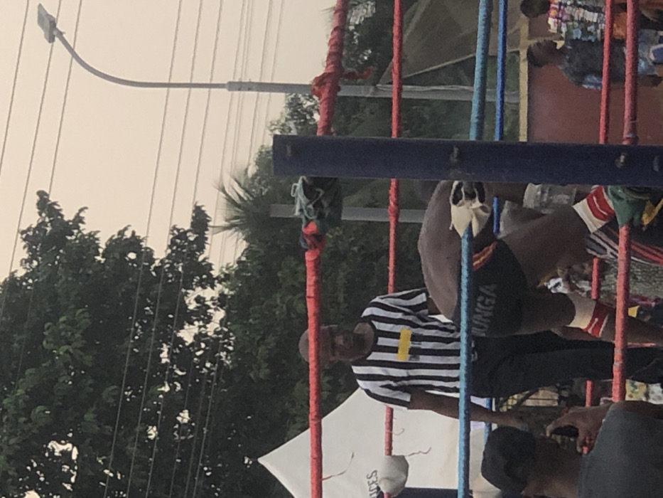 The scene depicts an outdoor professional wrestling event in Alor, Nigeria. In the foreground, a dark-skinned male wrestler, wearing black trunks with "JUNGA" printed in white on the rear, and red and white leg wraps, is bent over at the waist with his back to the viewer, positioned inside a wrestling ring defined by red and blue ropes and a prominent blue corner post. Behind him, a dark-skinned male referee in a black and white vertical striped shirt featuring a central horizontal yellow stripe and dark trousers stands facing into the ring. To the immediate left, a spectator in a dark shirt and black cap is visible from the shoulders up, holding a mobile phone in a recording or photographing stance. The background features lush green foliage from numerous trees, a partial view of a white tent on the left, multiple overhead power lines, and a tall street light with a curved pole on the right, all under an overcast or hazy sky.