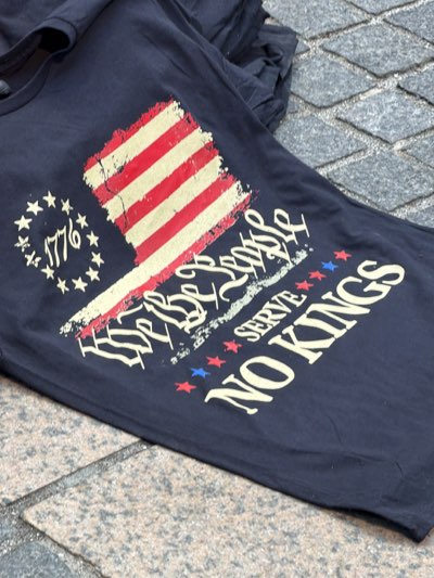 The image shows a black t-shirt lying on a gray stone surface. The t-shirt has a graphic print featuring a distressed American flag motif. To the left of the flag is a circle of stars with "1776" inside. Under the flag, the phrase "WE THE PEOPLE SERVE NO KINGS" is printed. The background suggests an outdoor setting, potentially a sidewalk or pavement. There is no evidence to link the image specifically to New York City.
