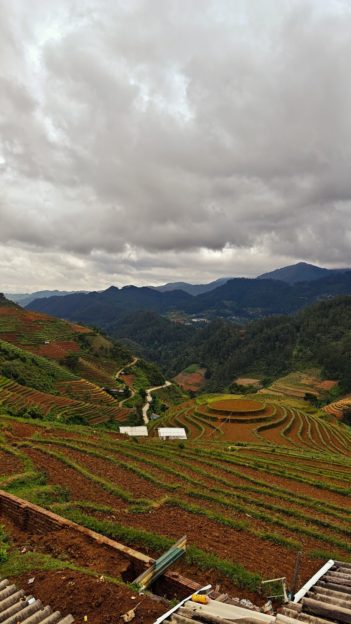 This vertical image depicts a panoramic view of terraced agricultural fields in a mountainous region of Dế Xu Phình, Vietnam, under an overcast sky. The upper third of the frame is dominated by a dense layer of grey, nimbostratus clouds, with brighter patches indicating diffused light. Below the sky, several layers of dark green, forested mountains recede into the distance, appearing hazy.
The midground showcases extensive terraced fields. These terraces vary in color, primarily reddish-brown exposed soil, with some areas exhibiting light green vegetation, suggesting early stages of crop growth. A winding, unpaved road snakes through the hillsides, visible amidst the terraces. Two distinct structures with white corrugated roofs are situated in the mid-left portion of the terraced landscape. Further in the valley, scattered small buildings can be discerned against the backdrop of the mountains. A prominent circular terraced formation, characterized by concentric rings of reddish-brown soil, is visible in the lower right-midground.
The immediate foreground reveals elements of a foreground structure or vantage point. This includes a section of a brick wall, disturbed earth, and corrugated roofing sheets. Debris, including a yellow can, is scattered on these surfaces, indicating an elevated, possibly residential or construction site from which the photograph was taken. No individuals are present within the frame.