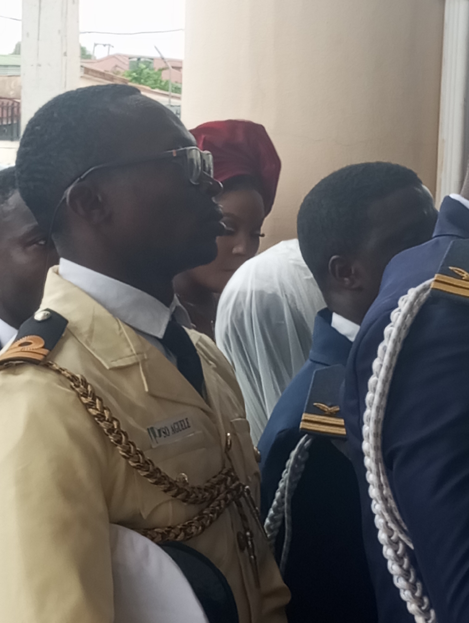 The image shows a close-up of several people in formal attire, possibly at an event in Barden Yana, Nigeria. In the foreground, a person wearing a light-colored uniform with gold braiding and epaulets with gold insignia is visible from the chest up. The uniform has a dark tie and a white collar. The name "VSO AGELLF" is visible on the uniform. To the right of this individual, another person is seen wearing a dark blue uniform with white braided cords and gold insignia on the epaulets. Further to the left and in the background, other individuals are partially visible. One person has a dark head covering, and another is seen wearing glasses. The background appears to be an interior or covered area with a pale wall and architectural elements suggesting a building, possibly a church or event hall, with hints of outdoor greenery and buildings visible through an opening. The lighting is diffused, indicating an overcast or indoor setting.