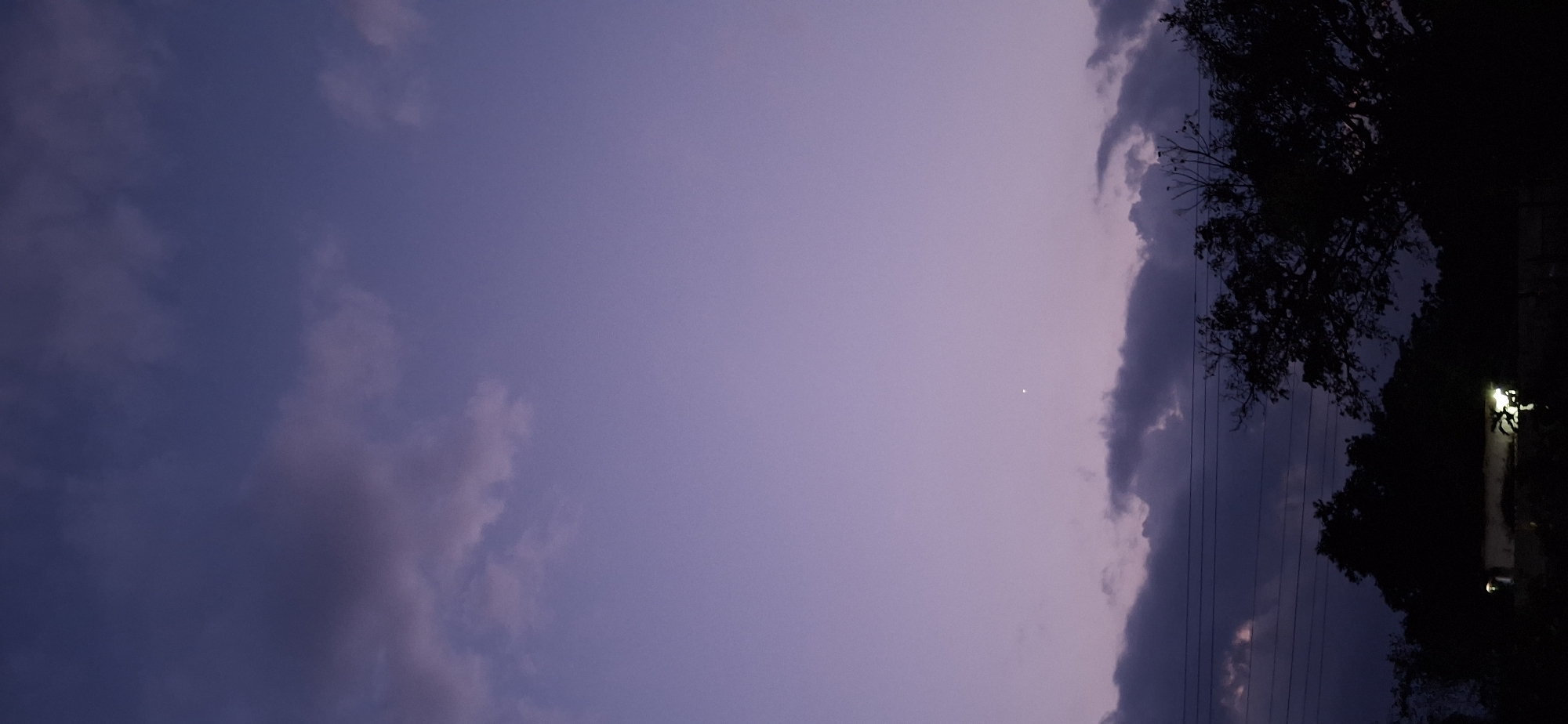 The image depicts a twilight sky with a range of colors from light purple to dark blue-grey, indicating sunset or twilight. The sky is mostly clear with some scattered clouds. A bright star is visible in the center-right of the image. On the right side, there is a silhouette of trees and what appears to be a streetlamp or light fixture. Power lines run vertically along the right side of the image. It is not possible to determine a specific location in Vallecito, Venezuela, from the image alone. No people or interactions are visible in the frame.