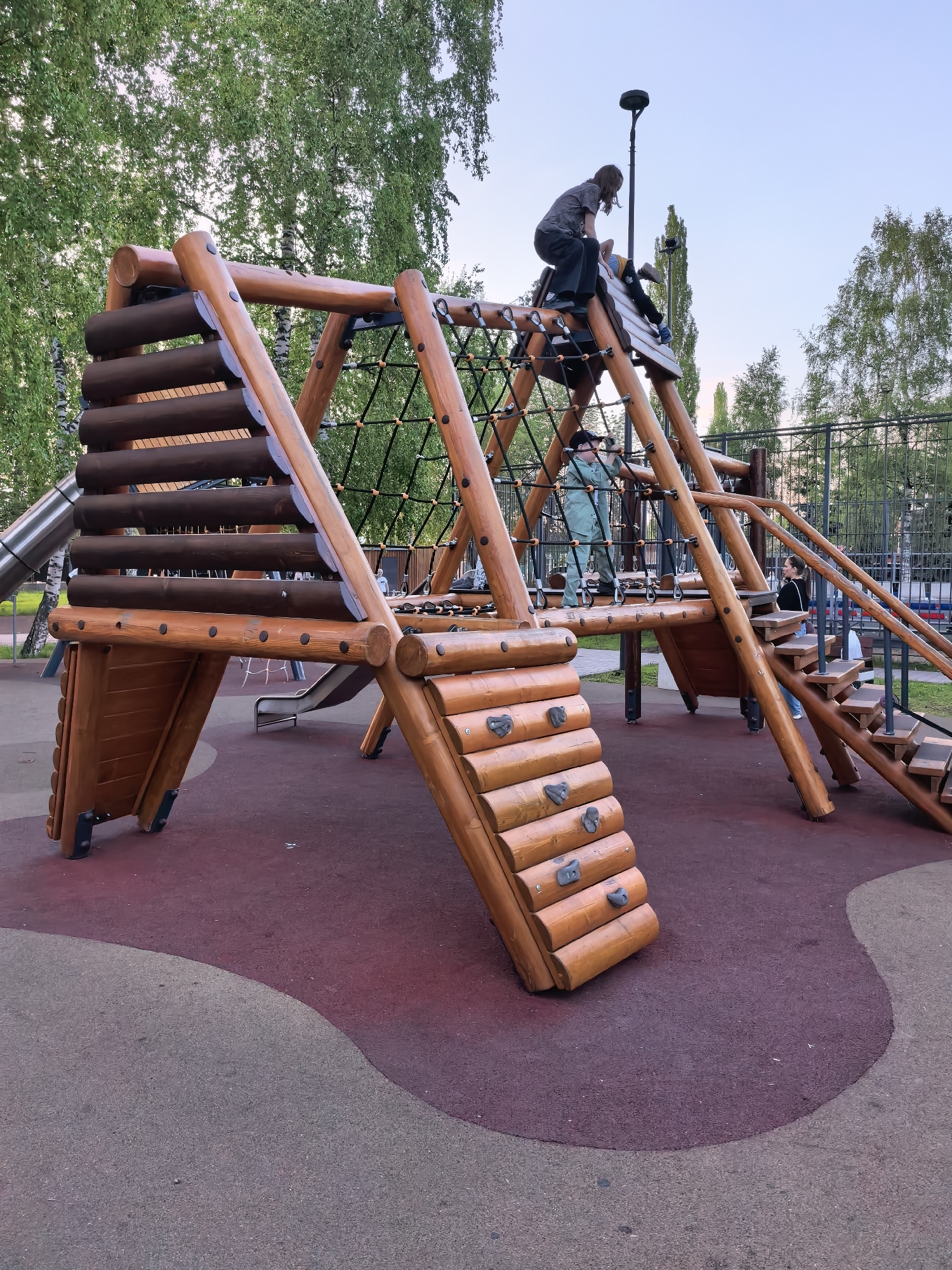 The image depicts a playground structure in Sterlitamak, Russia. The structure is made of wood and includes a climbing wall, stairs, a rope net, and a slide. Several children are climbing and playing on the structure. The playground is located in a park with trees and grass in the background. There is a fence bordering the playground. The ground is covered in a dark maroon and gray safety surface. A street lamp is visible behind the structure.