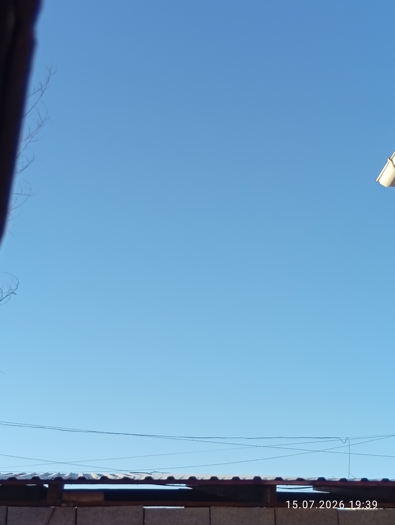A clear, light blue sky dominates the upper two-thirds of the image. In the top right corner, a white object, possibly a gutter or downspout, is visible against the sky. The lower third of the image shows the corrugated metal roof of a structure, supported by wooden beams. A wooden fence is visible beneath the roof. Thin, dark wires run horizontally across the sky above the roof. On the bottom right, a timestamp reads "15.07.2026 19:39". The scene appears to be outdoors in Ak-Suu, Kyrgyzstan.