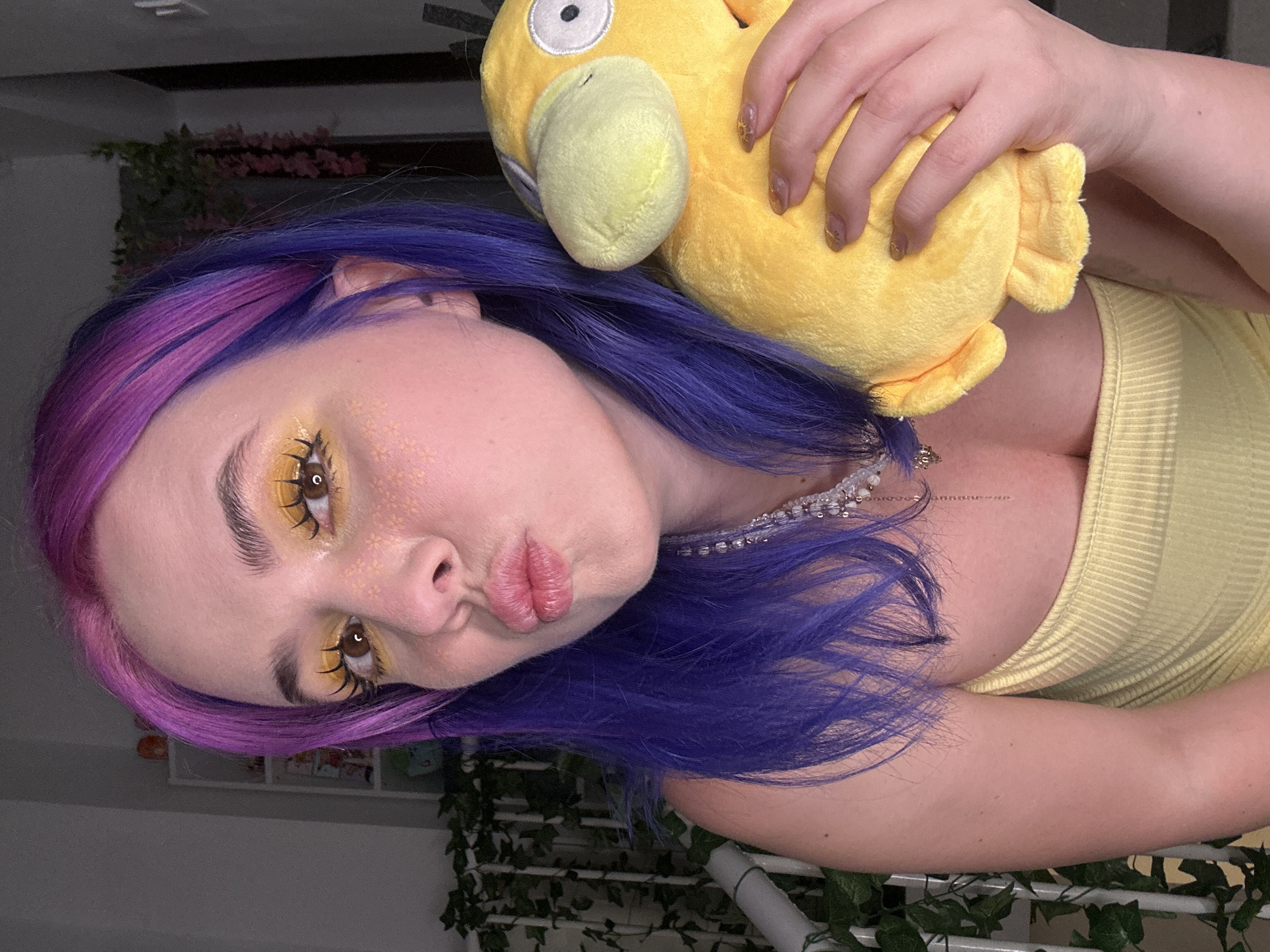 A person with purple and pink hair is holding a yellow Psyduck plush toy. They are wearing a yellow top and a pearl necklace. Their makeup includes yellow eyeshadow and glitter around the eyes. The background includes greenery and a shelf with items on it. The image appears to be taken indoors. The location is not discernible as Zagreb, Croatia based on the given image.